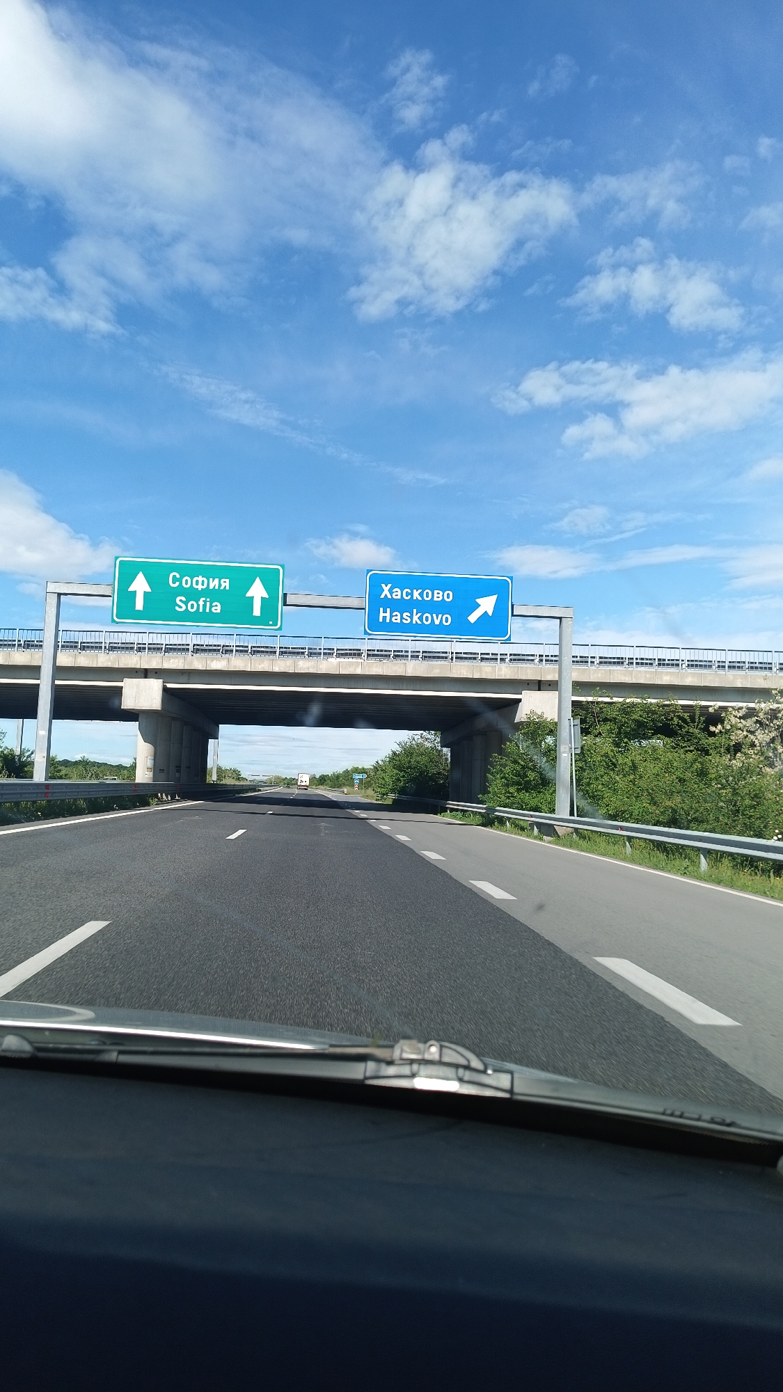 The image shows a view from inside a vehicle driving on a highway in Zagore, Bulgaria. Overhead signs indicate the routes to Sofia (straight ahead) and Haskovo (to the right). The highway is paved and marked with white lines. An overpass is visible in the distance. The sky is blue with scattered clouds. The environment appears to be rural with vegetation visible along the sides of the highway.
