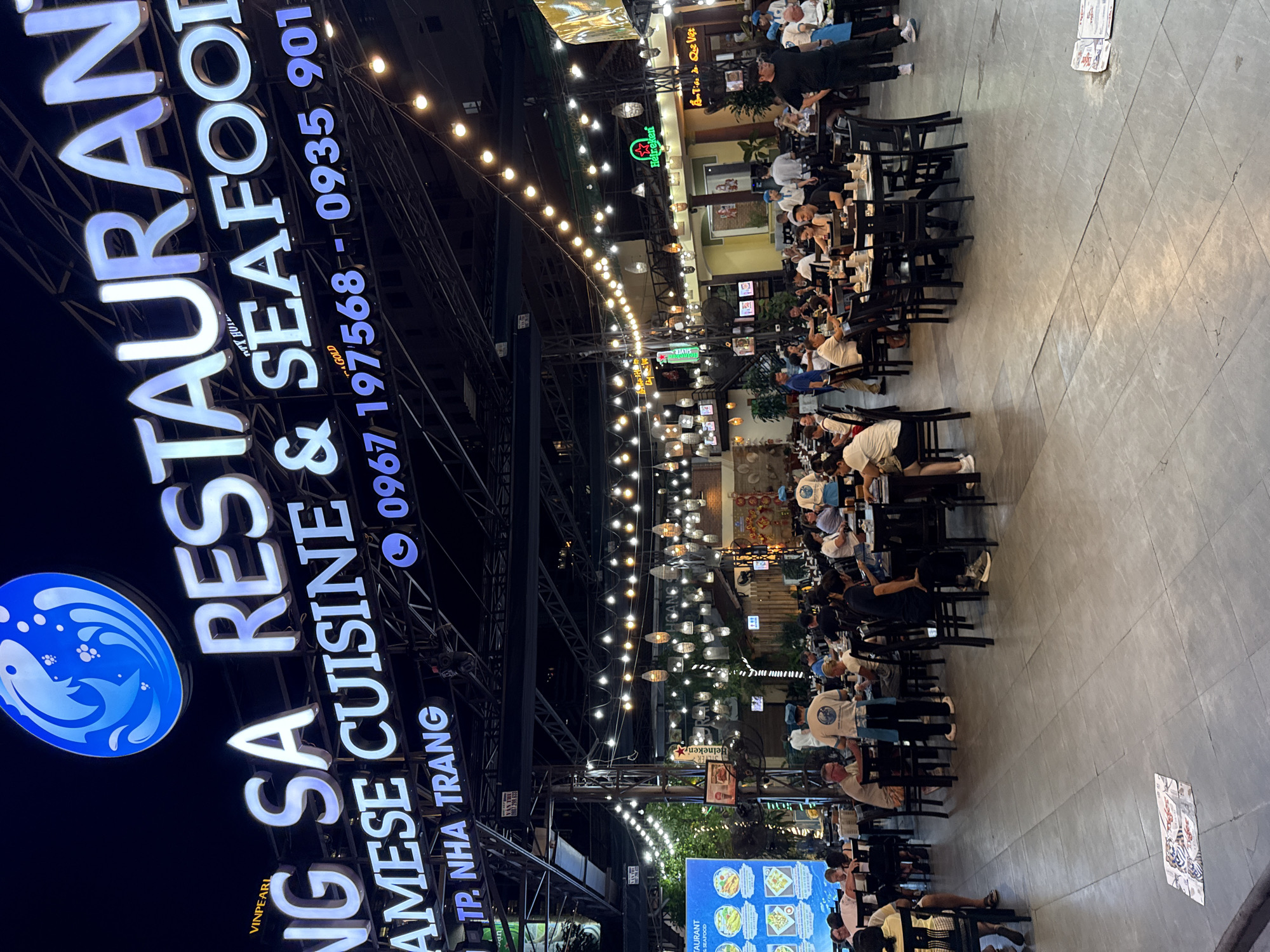 This is a nighttime, eye-level, medium shot of a busy outdoor restaurant in Nha Trang, Vietnam. The prominent signage for "NG SA RESTAURANT VIETNAMESE CUISINE & SEAFOOD" with a blue fish logo illuminates the scene. Below the main sign, contact information "0967 197568 - 0935 901" and "TP. NHA TRANG" are visible. Numerous string lights are draped across the open-air seating area, illuminating tables where patrons are dining. Several staff members in uniform are visible, including one in a white shirt with a blue apron, interacting with diners and clearing tables. The restaurant appears to be full with many people seated at tables, some eating, some talking, and some looking at their phones. In the background, more seating areas and building structures are visible, suggesting a large establishment. The floor is paved with large, grey tiles, and on the ground near the foreground, there are several printed placemats. The overall atmosphere is lively and social.