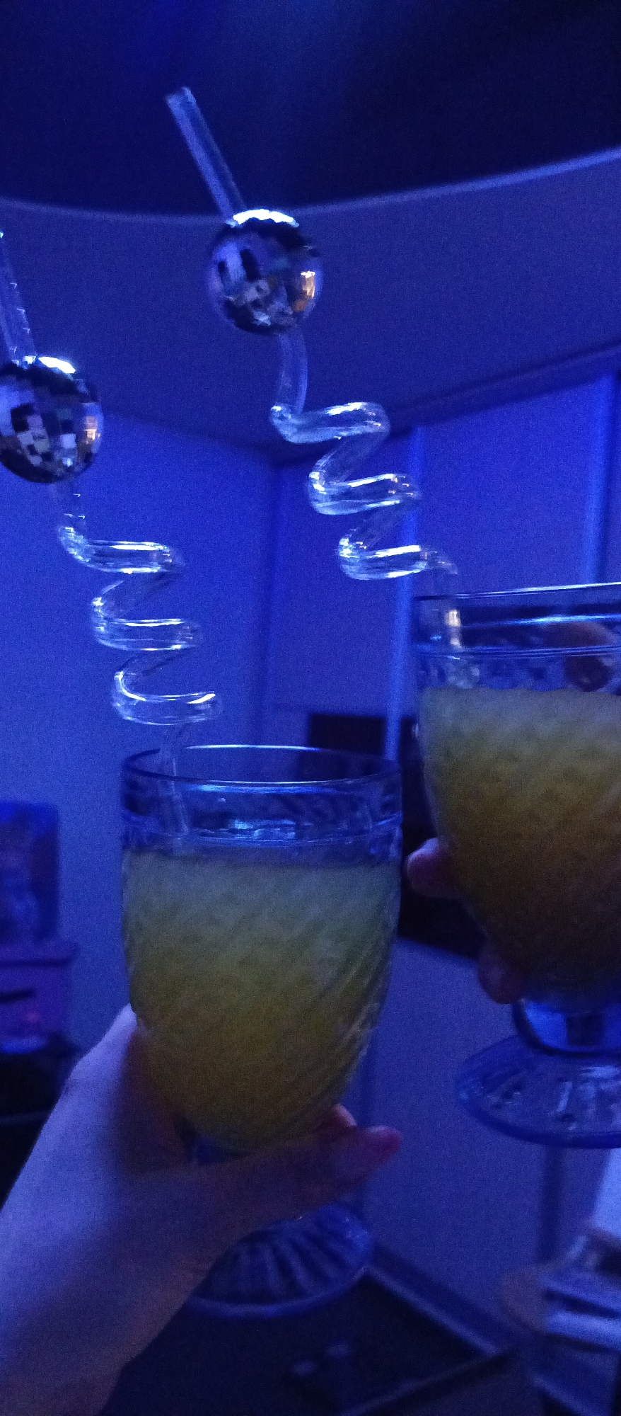 Two blue-tinted glasses filled with a yellow liquid are held up, presumably in a toast. Each glass has a clear, swirly straw, with a small disco ball attached to the top of each. The lighting is predominately blue, giving the scene a somewhat club-like atmosphere. The location is undetermined.