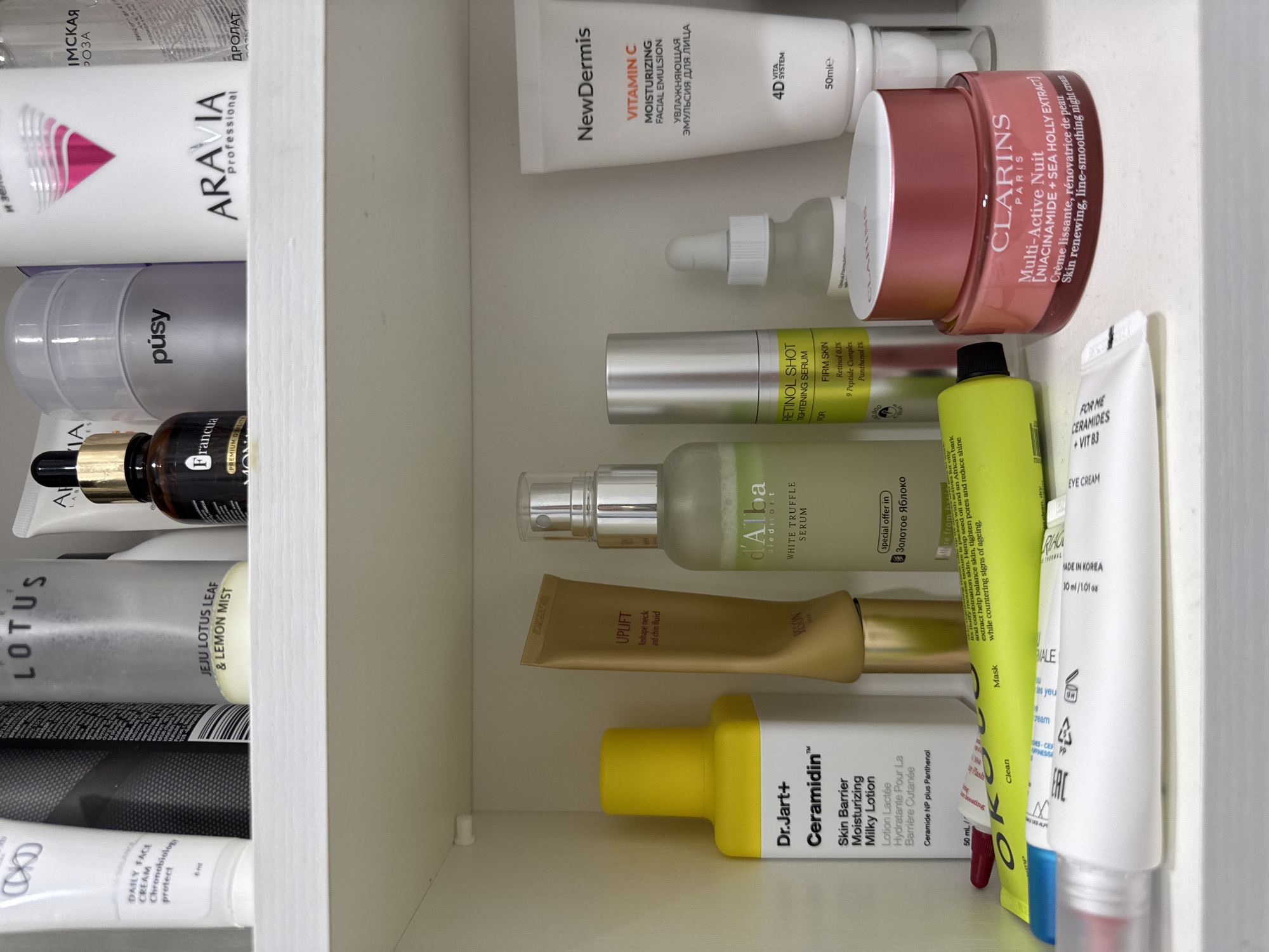 This is an indoor shot, taken from above, of a shelf filled with various skincare products. Some visible brands include Aravia, Pústy, Francua, Lotus, Dr. Jart+, d'Alba, Riag, NewDermis, and Clarins. The products are a mix of bottles and tubes, including lotions, serums, creams, and masks. The location, Sochi, Russia, is not evident from the image content.