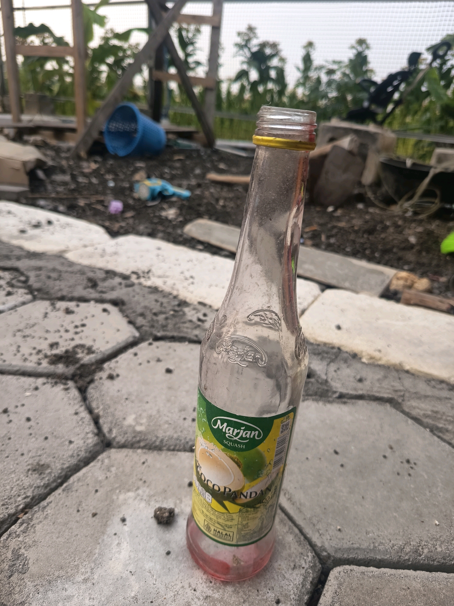In Sidorejo, Indonesia, an empty glass bottle of Marjan Squash, likely coconut pandan flavor, stands tilted on a textured concrete paver surface. The bottle, clear with a yellow cap and a green and yellow label, contains a small amount of pink liquid at the bottom. The label features text, including "Marjan," "SQUASH," "COCO PANDAN," and "400 ml," as well as a "HALAL" certification mark. The background is cluttered with debris, including a blue plastic basket, a small purple object, and other unidentifiable items, suggesting an outdoor, possibly neglected area. Wooden structures and out-of-focus greenery are visible further in the background, hinting at an outdoor construction or garden site. The lighting is overcast, casting a diffused light on the scene.
