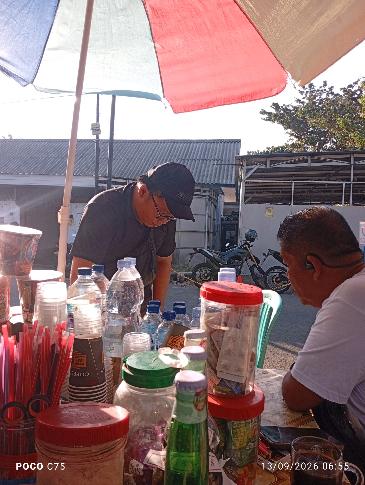 A middle-aged man wearing a black baseball cap, glasses, and a dark t-shirt is standing at a table with various items, including bottles of water, disposable cups, and straws. He is looking down at the table. In the background, another man in a white t-shirt and an earpiece is seated and facing away from the camera. A motorcycle is parked behind the table. The scene appears to be outdoors, possibly a street food stall, under a large umbrella that is red, white, and blue. The time stamp on the image indicates 13/09/2026 06:55. This scene is located in Jepara, Indonesia.