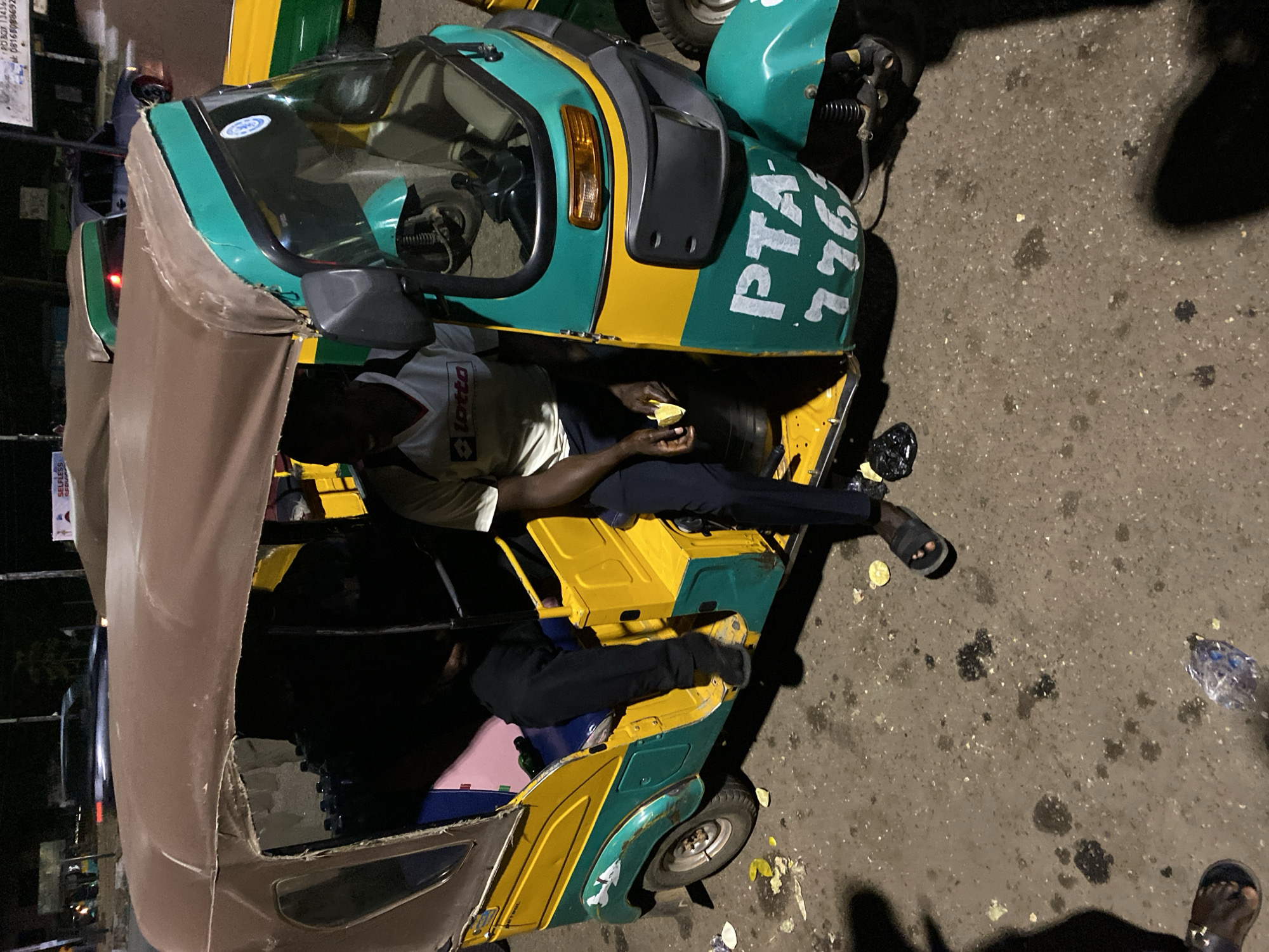 The image shows a three-wheeled vehicle, commonly known as a "Keke Napep" or auto-rickshaw, in Bukuru, Nigeria. The vehicle is primarily green and yellow with the inscription "PTA-776". A man in a "Lotto" shirt is sitting inside the vehicle, holding what appears to be a piece of fruit. A second person is in the back, wearing black pants. The vehicle's canvas roof is partially covering the side. The ground is paved and marked with dark stains. Additional feet are visible at the bottom. A sign post can be seen blurred in the background.
