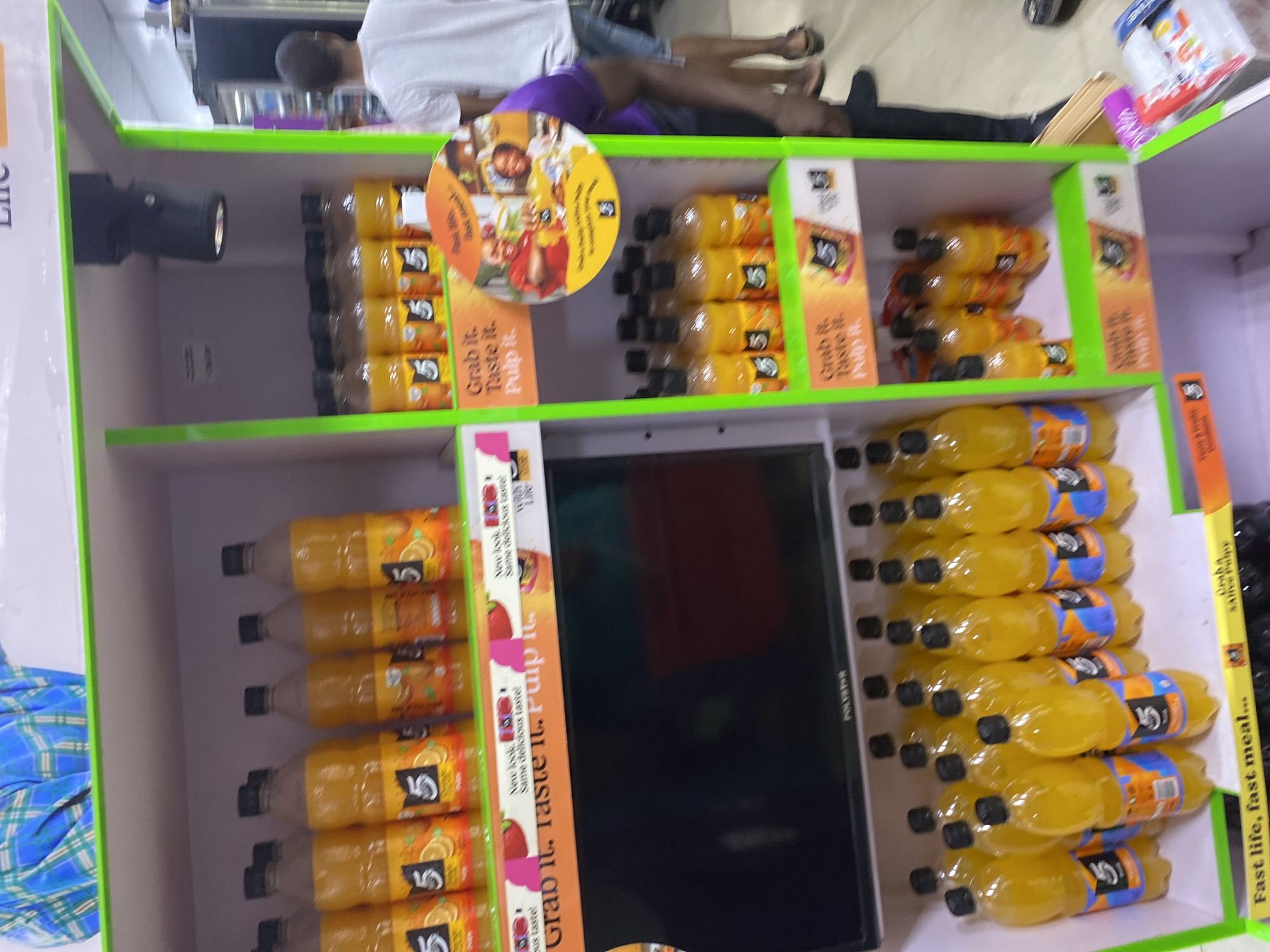 The image shows shelves stocked with bottles of "5 Alive" juice. The bottles are arranged neatly in rows on two shelves. Above the top shelf, there are people lying down, obscuring the top of the frame. In the middle of the shelves, there is a TV or monitor, seemingly integrated into the display. There is text near the juice which reads, "Grab it. Taste it. Pulp it." The last words of text says "Fast life, fast meal...". It is impossible to determine the location in the city of Lagos, Nigeria based on this single image.