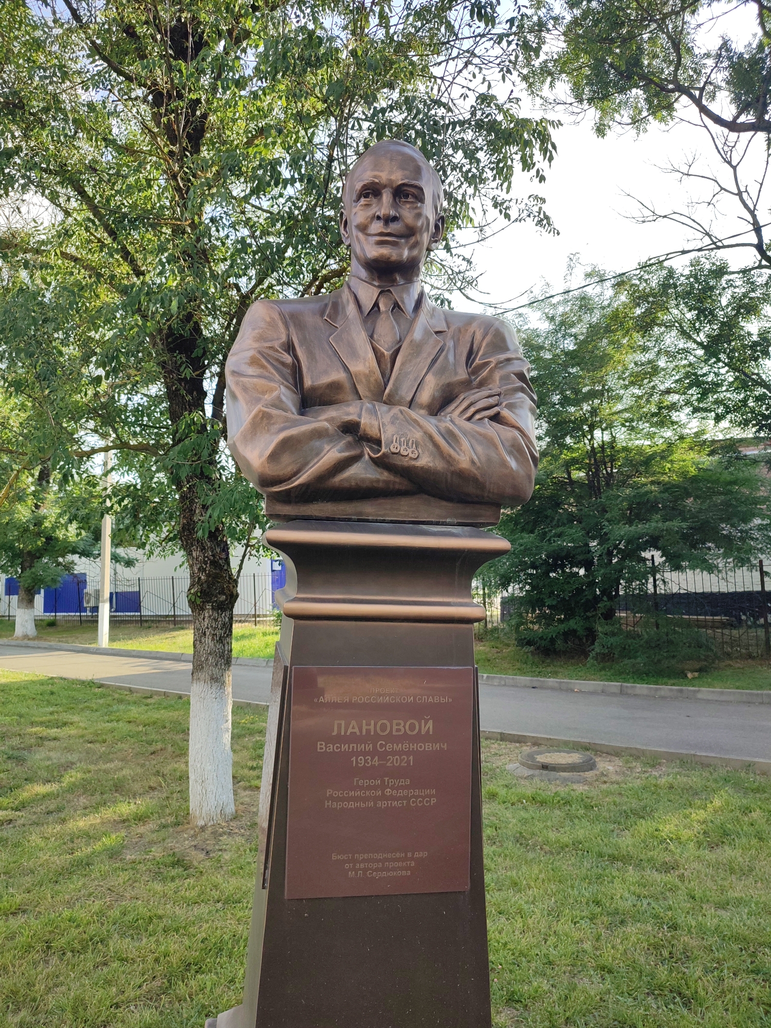 A bronze bust of a man, identified by an inscription as Vasily Semyonovich Lanovoy (1934-2021), a Hero of Labor of the Russian Federation and People's Artist of the USSR, stands on a pedestal outdoors. The pedestal has a plaque with this inscription, and also mentions it is part of the "Alley of Russian Glory" project. The bust depicts the man from the shoulders up, wearing a suit and tie, with his arms crossed. He has a stern expression. The bust is located in a grassy area with trees and a paved path in the background. Buildings and a fence are visible behind the trees. The environment appears to be a public space, possibly a park or memorial.
