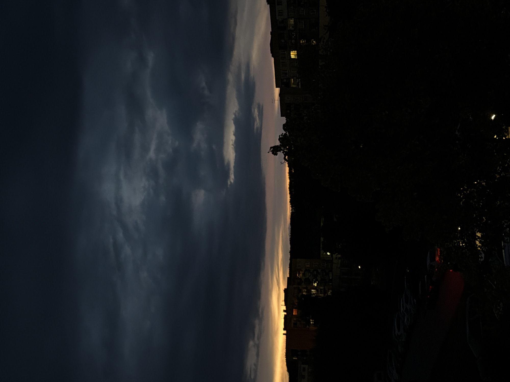 The image presents a nighttime view in an urban environment. The sky is dominated by heavy, dark storm clouds on the left, contrasting with a strip of lighter sky near the horizon, suggestive of either dawn or dusk. Buildings are visible along the horizon, some with illuminated windows, indicating human habitation. The right side of the image is shrouded in darkness, with trees and what appears to be parked vehicles discernible as silhouettes. A red taillight is visible among the parked vehicles. The orientation of the image is rotated ninety degrees counter-clockwise.