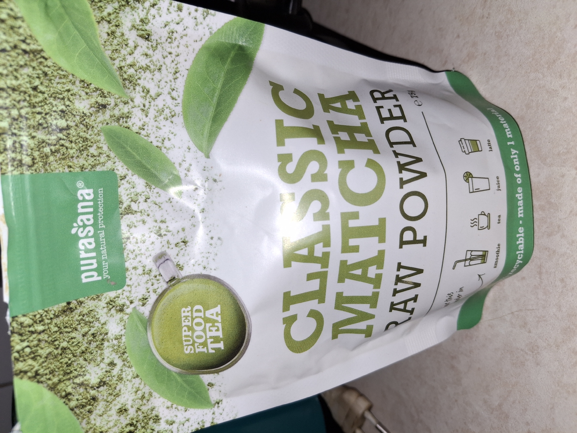 A close-up, eye-level, slightly angled shot shows a bag of Purasana Classic Matcha Raw Powder. The bag is predominantly white with green accents and text. The top half of the bag features a green band with the Purasana logo and "your natural protection" in white text. Behind this band, there's a texture resembling matcha powder interspersed with green leaves. A circular green graphic in the upper left reads "SUPER FOOD TEA." Prominently displayed across the middle of the bag in large, bold green lettering is "CLASSIC MATCHA RAW POWDER." Below this, the text "e 75g" is visible. The bottom portion of the bag shows small icons representing different ways to use the matcha: a smoothie, tea, juice, and latte. Text beneath these icons reads "recyclable - made of only 1 material." The bag is placed on a light-colored, textured surface that appears to be a countertop. The background is dark and out of focus. The image does not contain any specific elements indicating a location in Åsgårdstrand, Norway.