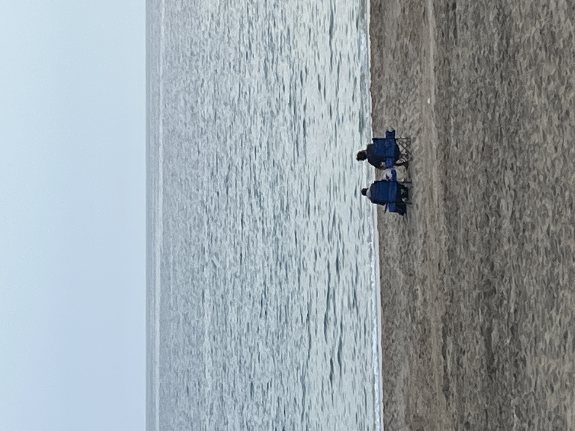The image shows two individuals sitting in blue beach chairs on a sandy beach next to a body of water. The water appears choppy with a greyish hue, possibly due to the weather. The scene is likely in a coastal area, but there is no identifiable landmark to determine the exact location in Alanya, Turkey, based solely on the image. The focus is on the two figures relaxing in their chairs near the water's edge.
