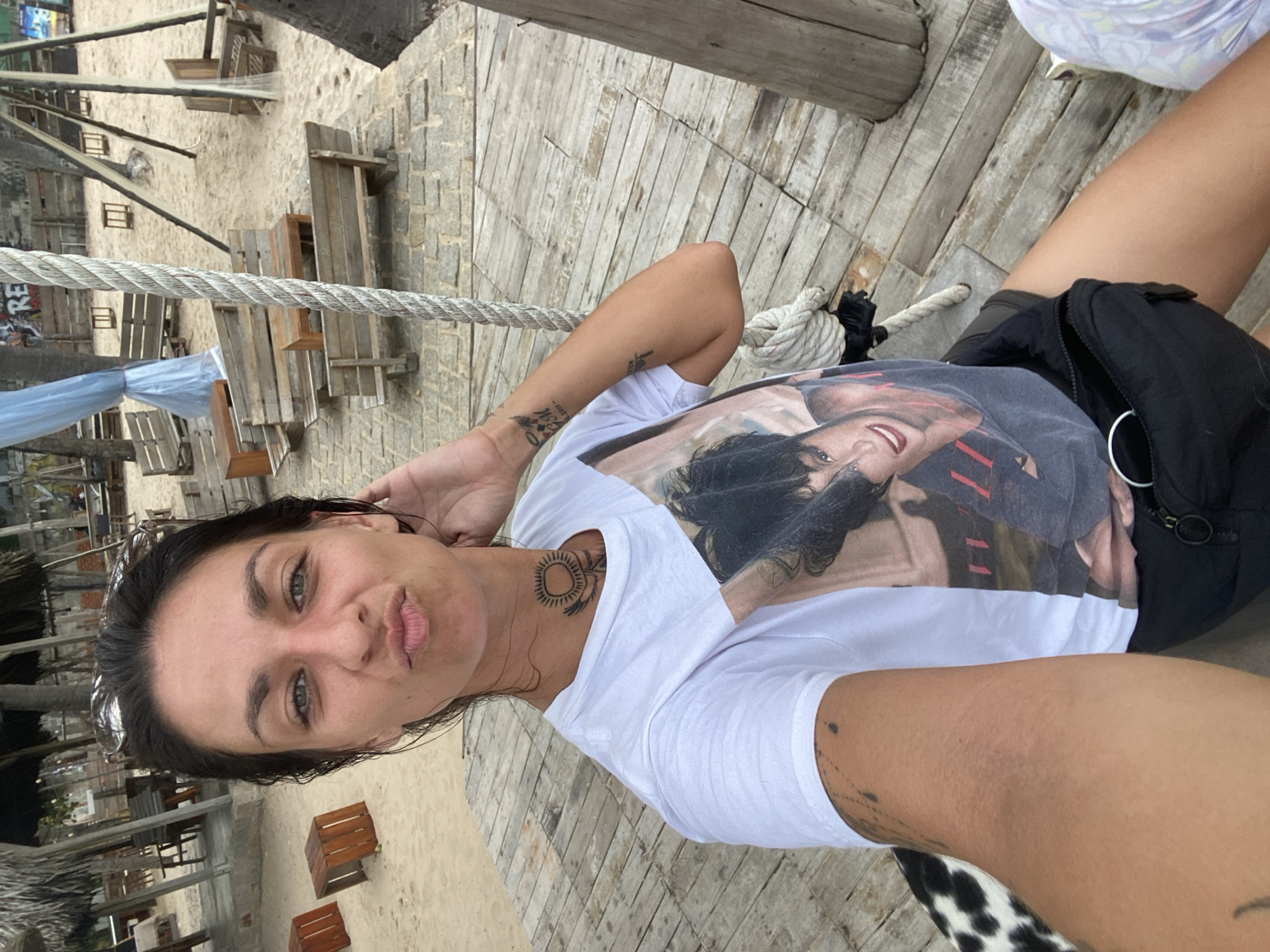 In Mũi Né, Vietnam, a woman is seated on a wooden swing on a pier with a sandy beach and thatched-roof structures in the background. She is wearing a white t-shirt with a graphic print, black shorts, and a fanny pack. The woman has dark hair, green eyes, and tattoos on her neck and arm. The scene depicts a relaxed, outdoor setting, likely a beachside resort or restaurant, with various wooden benches and tables scattered on the sand. The rope of the swing is prominent in the foreground, suggesting the woman is in the process of swinging or has just finished. The lighting indicates it is daytime, possibly overcast or late afternoon, given the soft shadows.