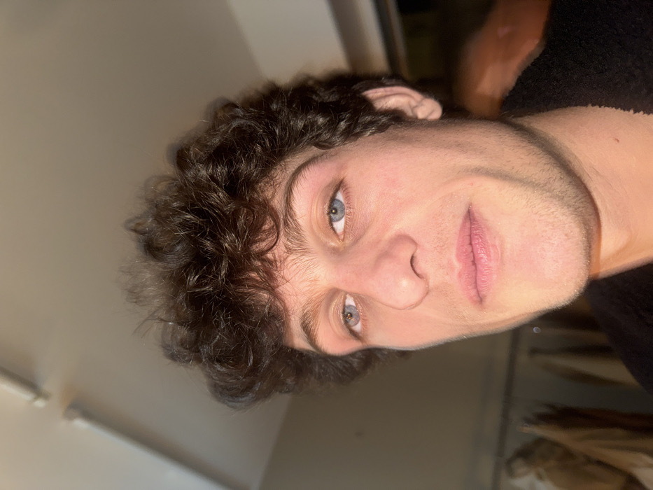 The image presents a close-up portrait of a male individual, positioned slightly off-center to the right. He has dark, curly hair, light blue eyes, fair skin, and visible stubble. The subject is looking directly into the camera. A dark, textured garment, possibly a fleece, is visible around his lower neck and shoulder area. The background is an out-of-focus indoor environment. A white linear light fixture is mounted on a light-colored ceiling in the upper left. To the subject's left, blurred shapes of light-colored hanging garments, likely on a clothing rack, are discernible. No specific external features or landmarks are visible to determine a precise location within the city of Paris, France; the scene is an indistinct indoor setting.