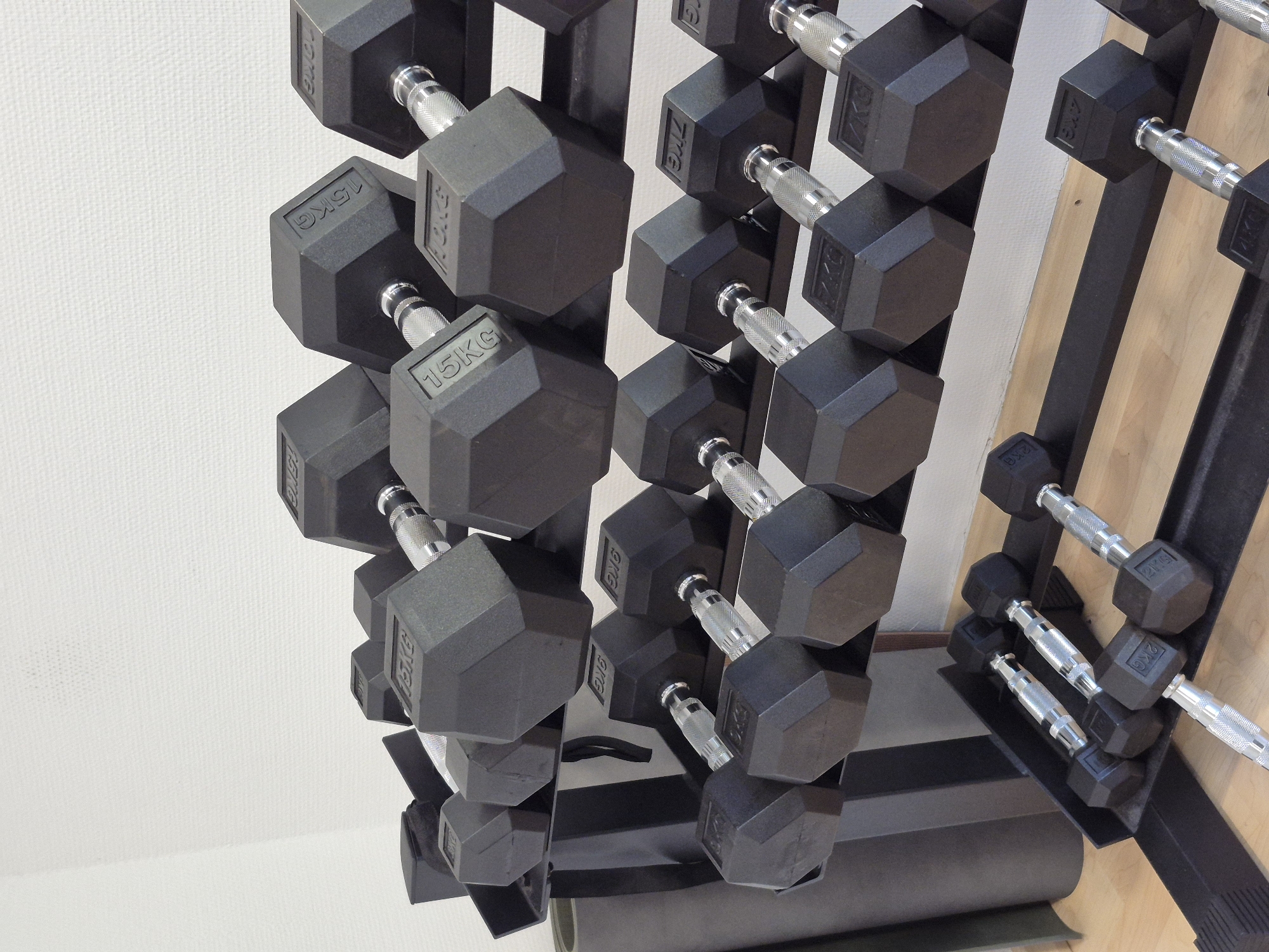 The image shows a rack of hexagonal dumbbells of various weights. The dumbbells are black with silver handles and have their weights clearly marked in kilograms. In the foreground, the rack holds dumbbells weighing 2kg and 4kg. In the middle row, dumbbells weighing 7kg are visible. The top row features heavier dumbbells, including ones marked as 10kg and 15kg. A dark grey exercise mat is leaning against the rack on the left side of the image. The dumbbells are arranged in an organized manner on a black metal rack. The background consists of a white, textured wall. The floor is made of light-colored wooden planks. The scene appears to be in a gym or fitness area within Schwäbisch Gmünd, Germany. No people are present in the image.