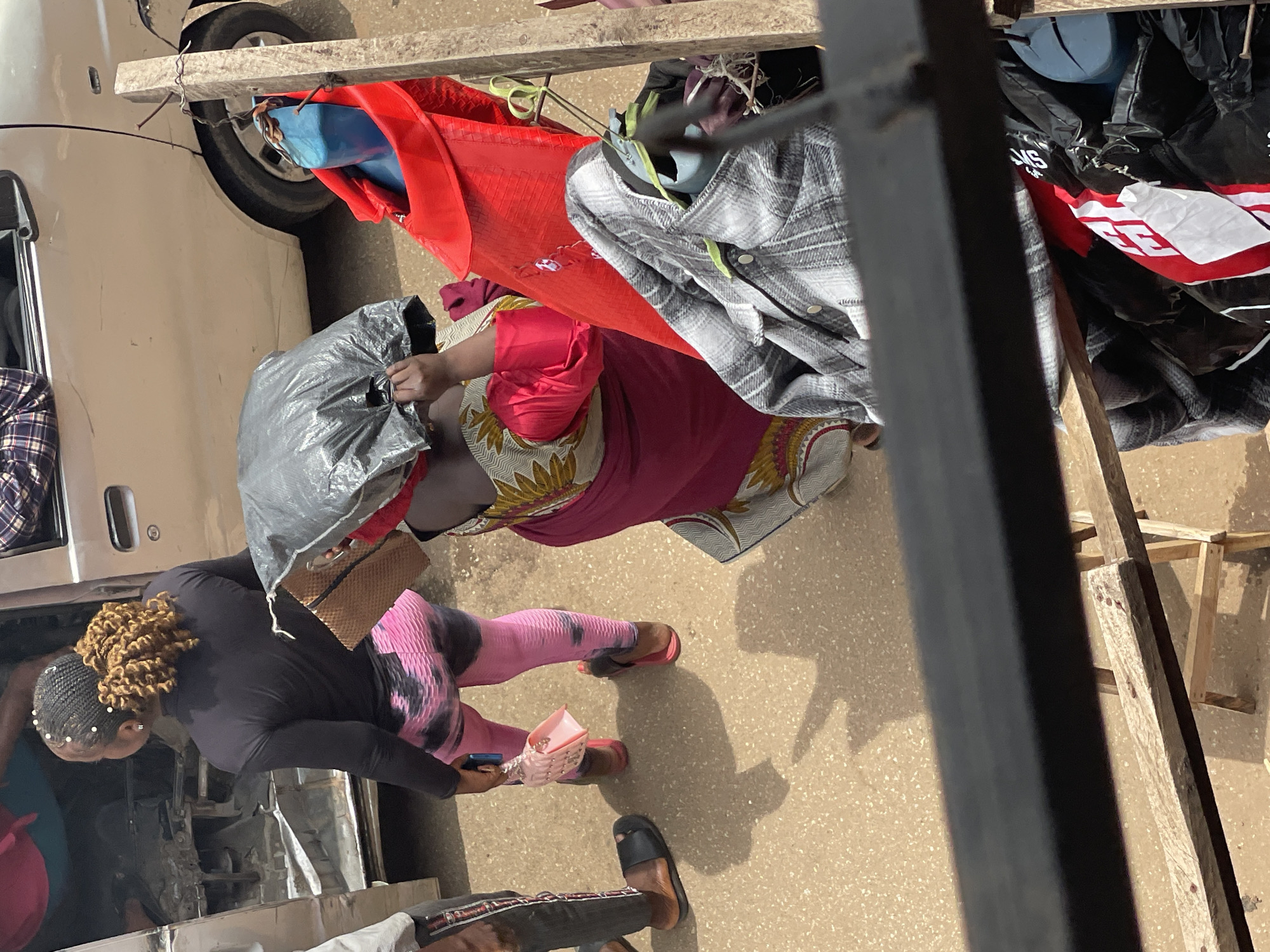 In an outdoor urban environment, three women are visible, two standing and one possibly seated within a vehicle. A woman in the foreground, facing away from the camera, wears a black long-sleeved top and pink tie-dye patterned leggings. She carries a small pink handbag and a large gray plastic bag. Another woman, partially visible on the right, is dressed in a vibrant pink and gold patterned garment. A third individual, mostly obscured, is seen on the left, wearing dark pants and a black sandal. They appear to be near a vehicle, possibly a van or truck, with its door open. The scene is characterized by a sandy or dirt ground surface and various items, including clothing and plastic bags, are scattered around. A wooden structure with a metal railing is also present in the foreground.