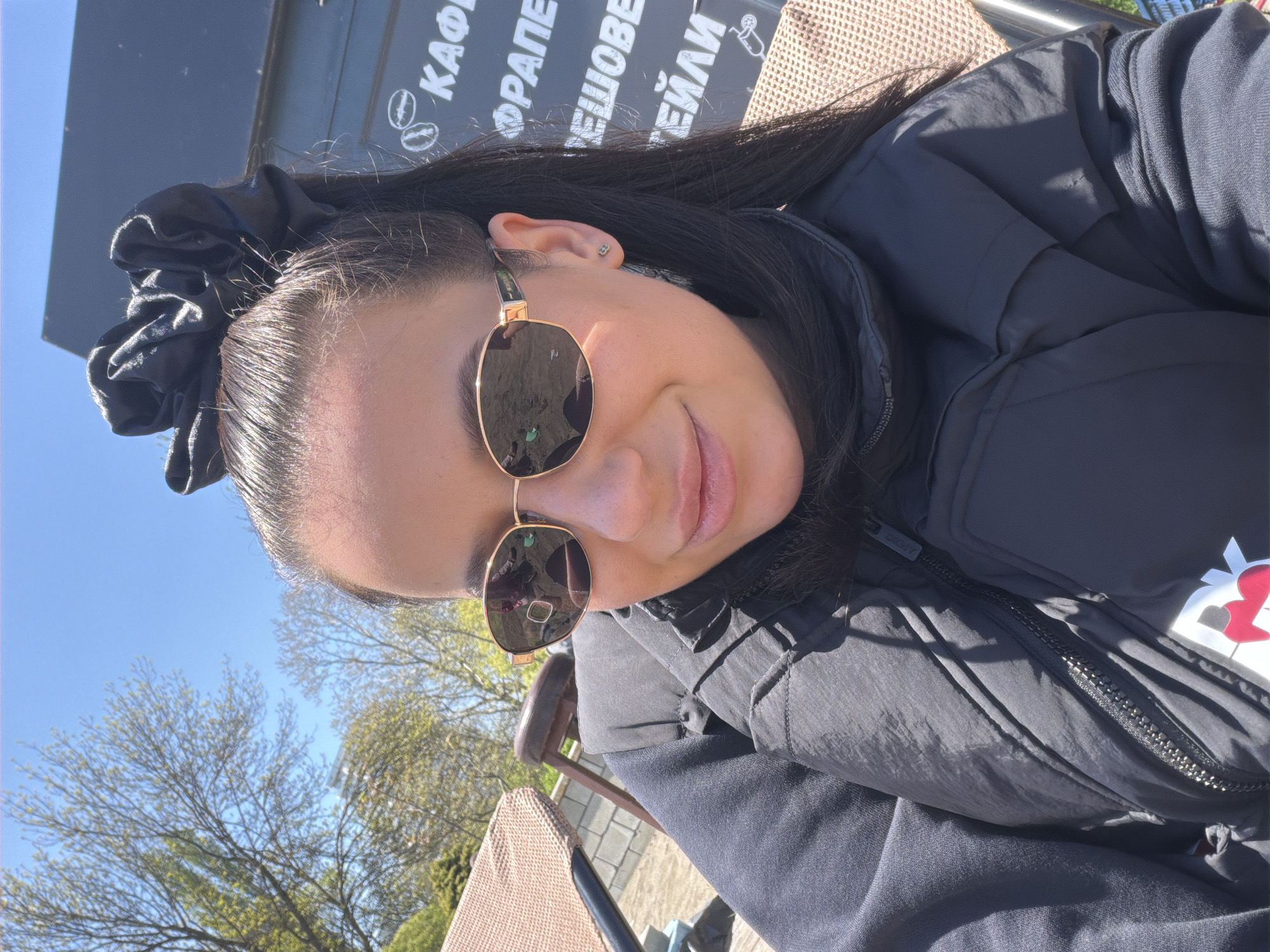 A woman is looking at the camera wearing sunglasses and a black jacket. She is sitting in a chair outdoors, with trees visible in the background. Behind her is a sign with writing in a Cyrillic alphabet. The location is Unknown, Unknown.