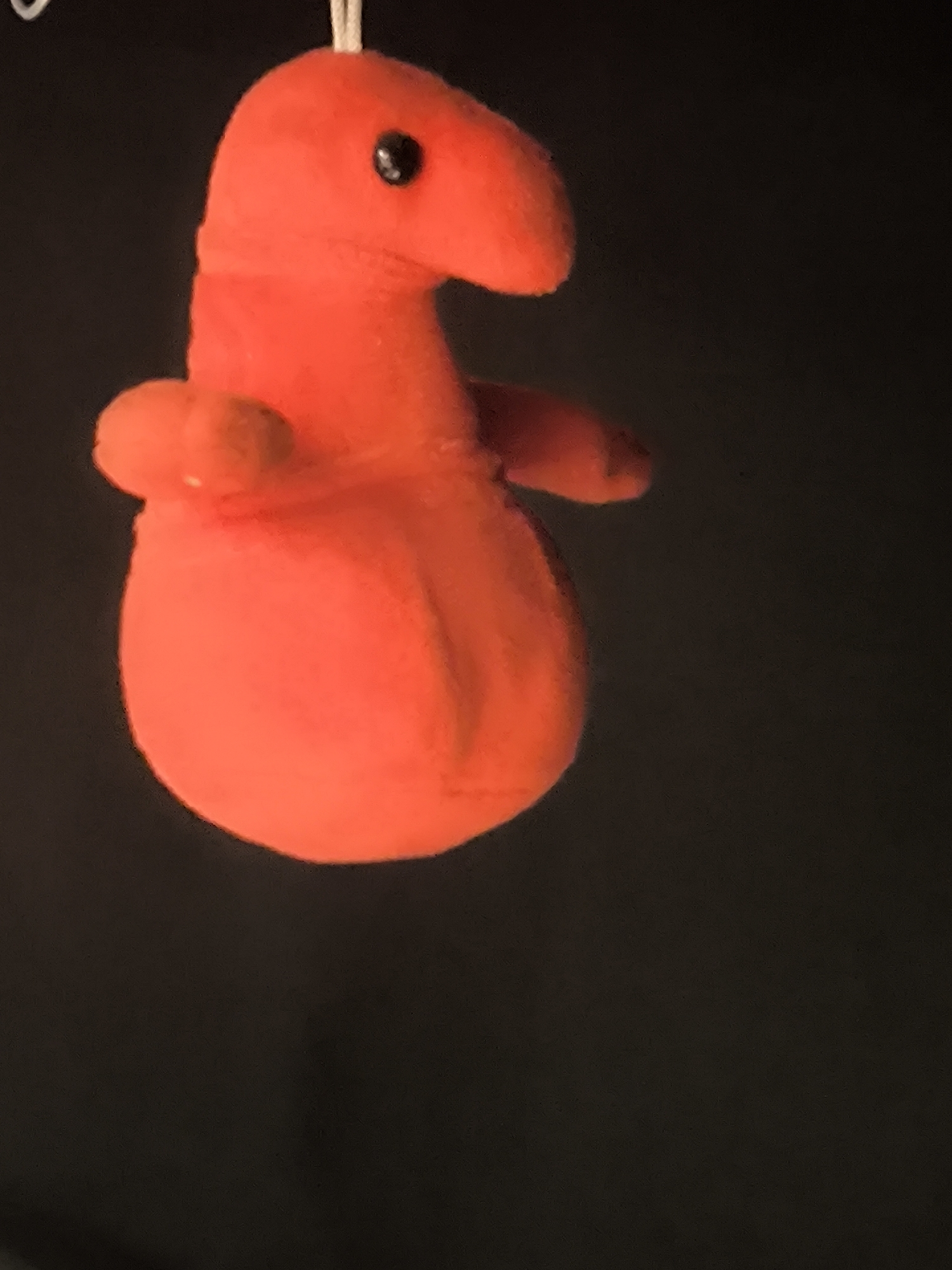 A plush toy, shaped like a red creature with a bulbous body and a long, pointed snout, is hanging against a black background. The toy has a small black eye visible on its snout and two small appendages resembling arms. The lighting appears to be from the side, casting some shadows and highlighting the texture of the plush material. The toy is suspended by a thin white string that is partially visible at the top of the image. There is no identifiable context of Otomin, Poland in the provided image.