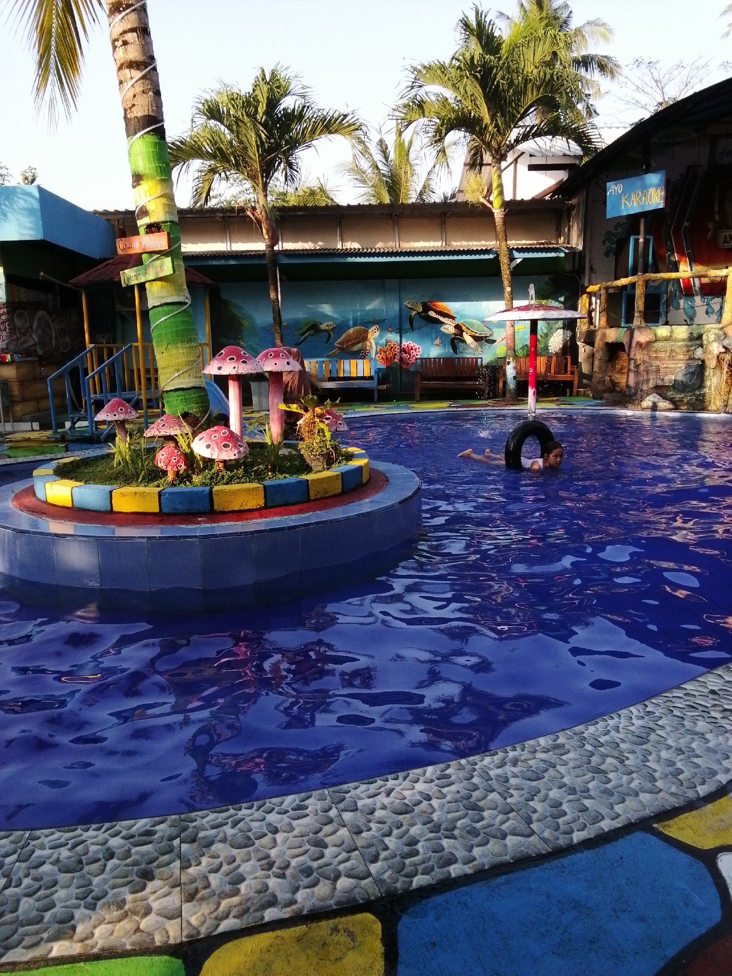A child swims in a blue swimming pool at the "Aqua Fantasy" water park in Kebonsari, Indonesia. The pool is surrounded by a colorful tiled edge and a gravel walkway. In the background, there is a large palm tree painted with green and yellow stripes, a mural of turtles and coral, and a building with a sign that reads "AYO KARAOKE." A few mushroom-shaped decorations are placed in a planter in the center of the pool. The water park appears to be outdoors, with a clear sky above.