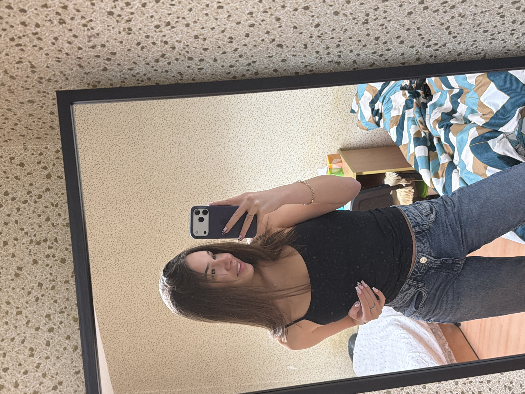 A woman is taking a selfie in a mirror. She is wearing a black tank top and blue jeans. Her nails are painted with a black and white print. She is smiling. The background of the photo shows a beige patterned wall, a bed with a blue and brown patterned comforter, a wooden nightstand, and a white blanket. The location appears to be indoors, possibly a bedroom. There is no evidence to suggest the location is in Sofia, Bulgaria.