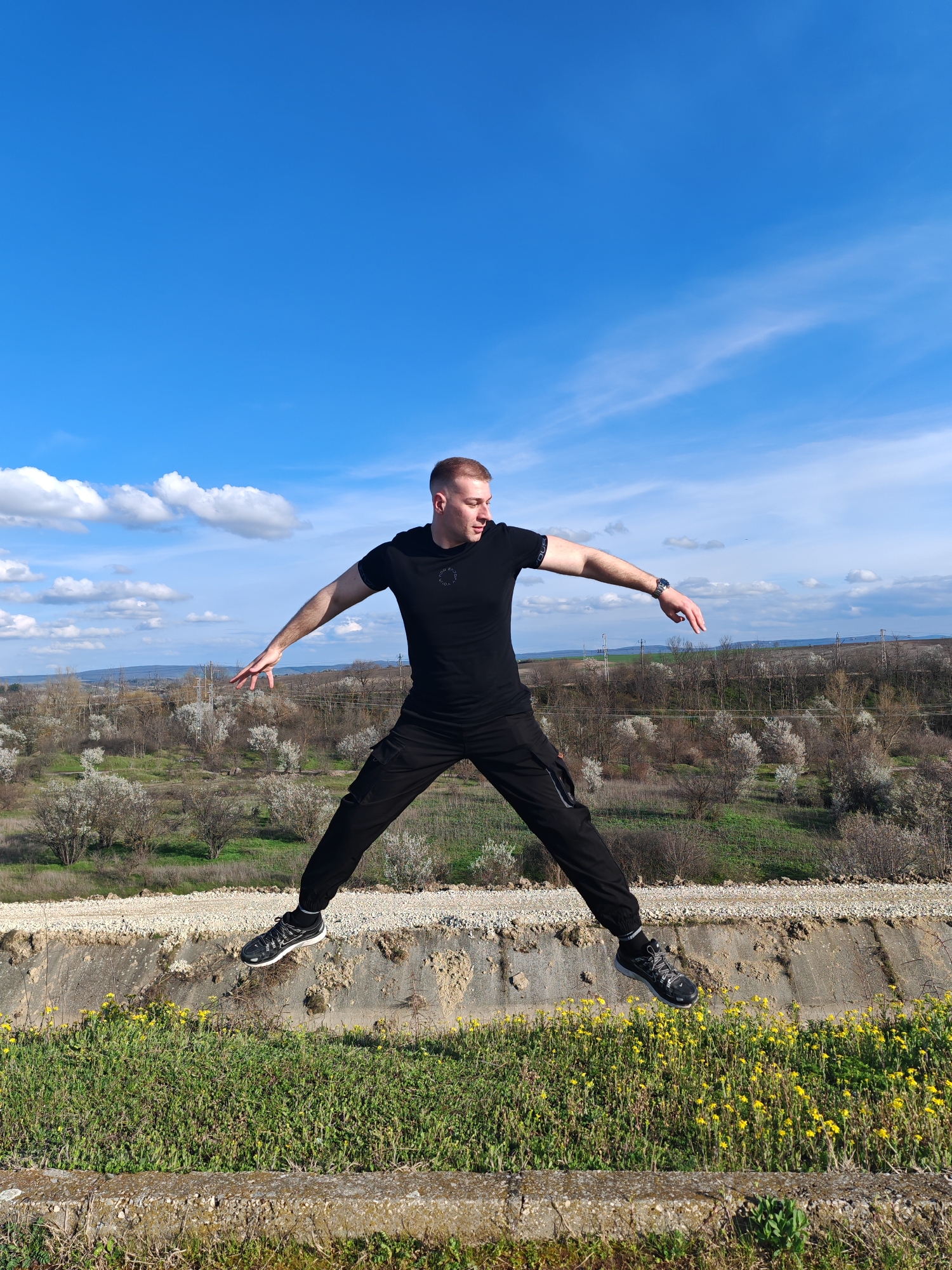 A man is jumping in the air on a grassy and concrete embankment, possibly a retaining wall or drainage feature. He is wearing black clothing, including a t-shirt and pants, and athletic shoes. His arms and legs are spread out in a star shape. Behind him is a field with blooming trees and a blue sky with scattered clouds. The location is in the city of Shumen, Bulgaria.