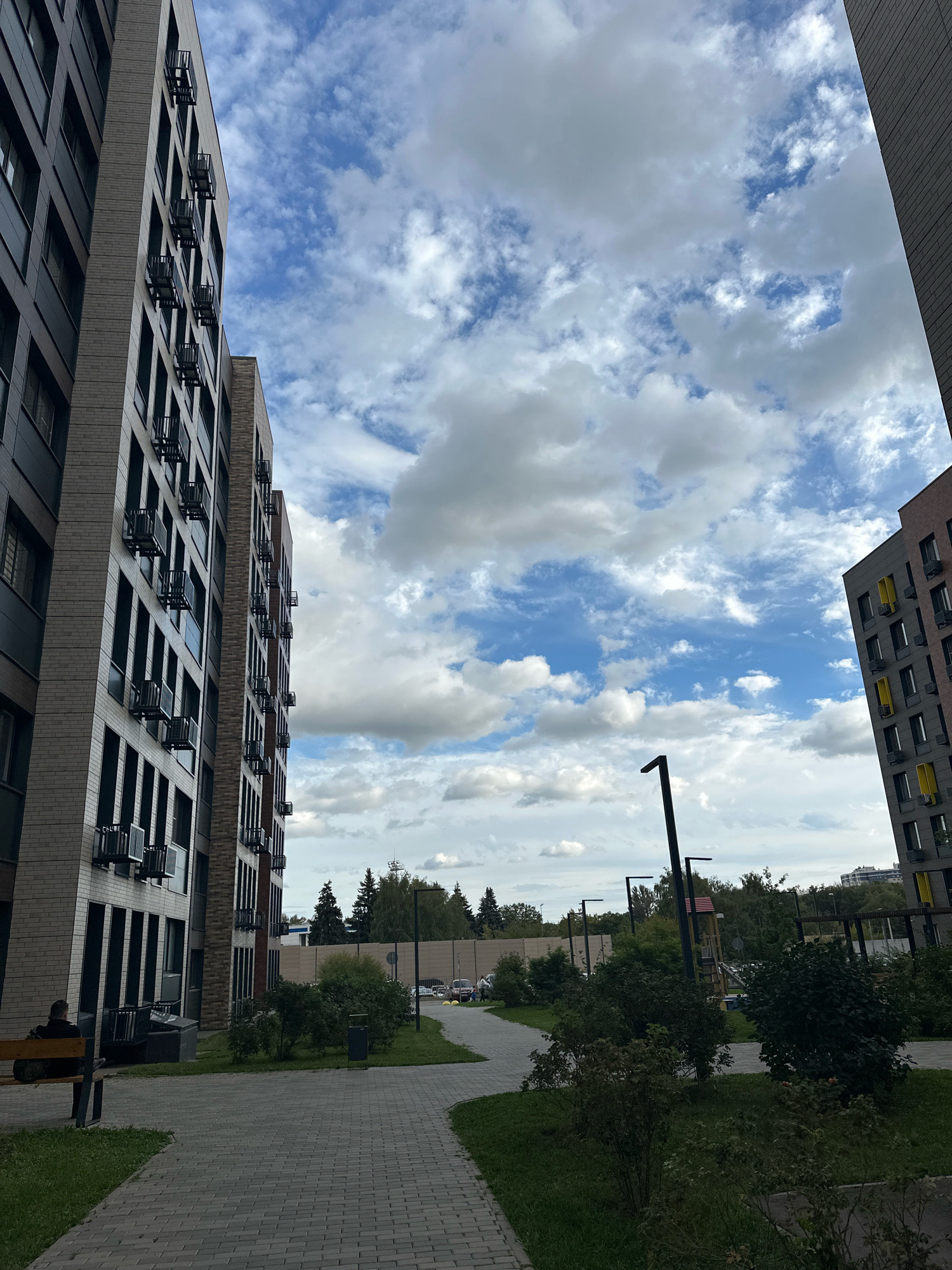 The image depicts an outdoor courtyard area with two modern, multi-story apartment buildings on either side. The building on the left is taller and features a facade of light brown brick and dark gray paneling, with numerous windows and external air conditioning units. The building on the right has a similar architectural style, with a dark gray facade punctuated by yellow accents around some windows, also equipped with external air conditioning units. A paved walkway with a brick pattern curves through the courtyard, bordered by grass and landscaped bushes. A single person is seated on a dark-colored bench on the left side of the frame, facing away from the camera. In the background, there are trees, a fence, and a glimpse of a playground structure. The sky above is a mix of blue sky and scattered white clouds. The scene appears to be in a residential urban environment, likely Vidnoye, Russia, given the architectural style and the presence of modern apartment complexes.