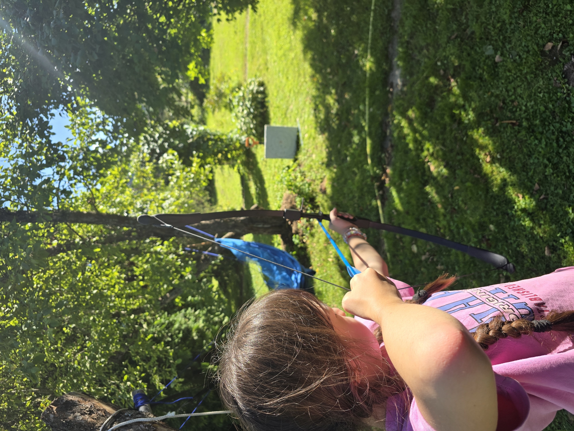 In the village of Lobkovo, Russia, on a bright, sunny day, a young girl with brown hair braided into a pigtail is practicing archery. She is positioned in a backyard with lush green grass and trees, with a target visible in the background. The girl wears a pink t-shirt with lettering on it. She is holding a bow and arrow, aiming at the target, her body angled towards it. The scene appears to be set up for a recreational activity, with a blue hammock visible in the background, adding to the casual outdoor setting.