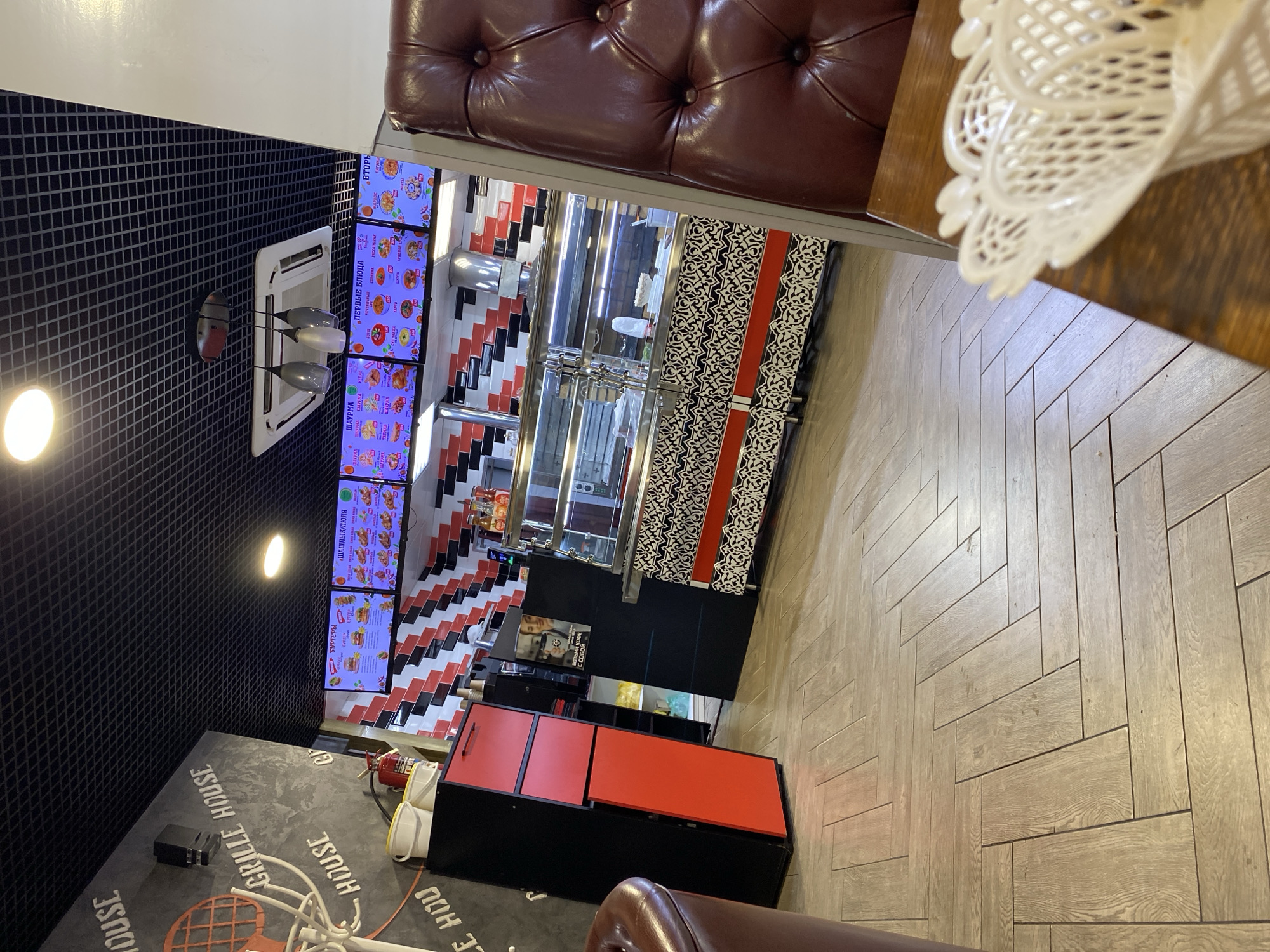 The image captures the interior of a fast-food restaurant, likely a "Grill House" as suggested by the signage on the wall. The restaurant features a modern, somewhat industrial design with a black grid ceiling and tiled walls in red, black, and white. Digital menus displaying food items like burgers and shashlik are prominently displayed on screens above a food display counter. The counter itself contains a glass display case, showing food items, and a stainless steel exhaust hood. In the foreground, a red leather booth seat is partially visible, and a white plastic basket sits on a wooden table. The flooring is a light-colored wood-look tile laid in a herringbone pattern. The overall impression is of a clean and functional dining space. The location is identified as Ishimbay, Russia.