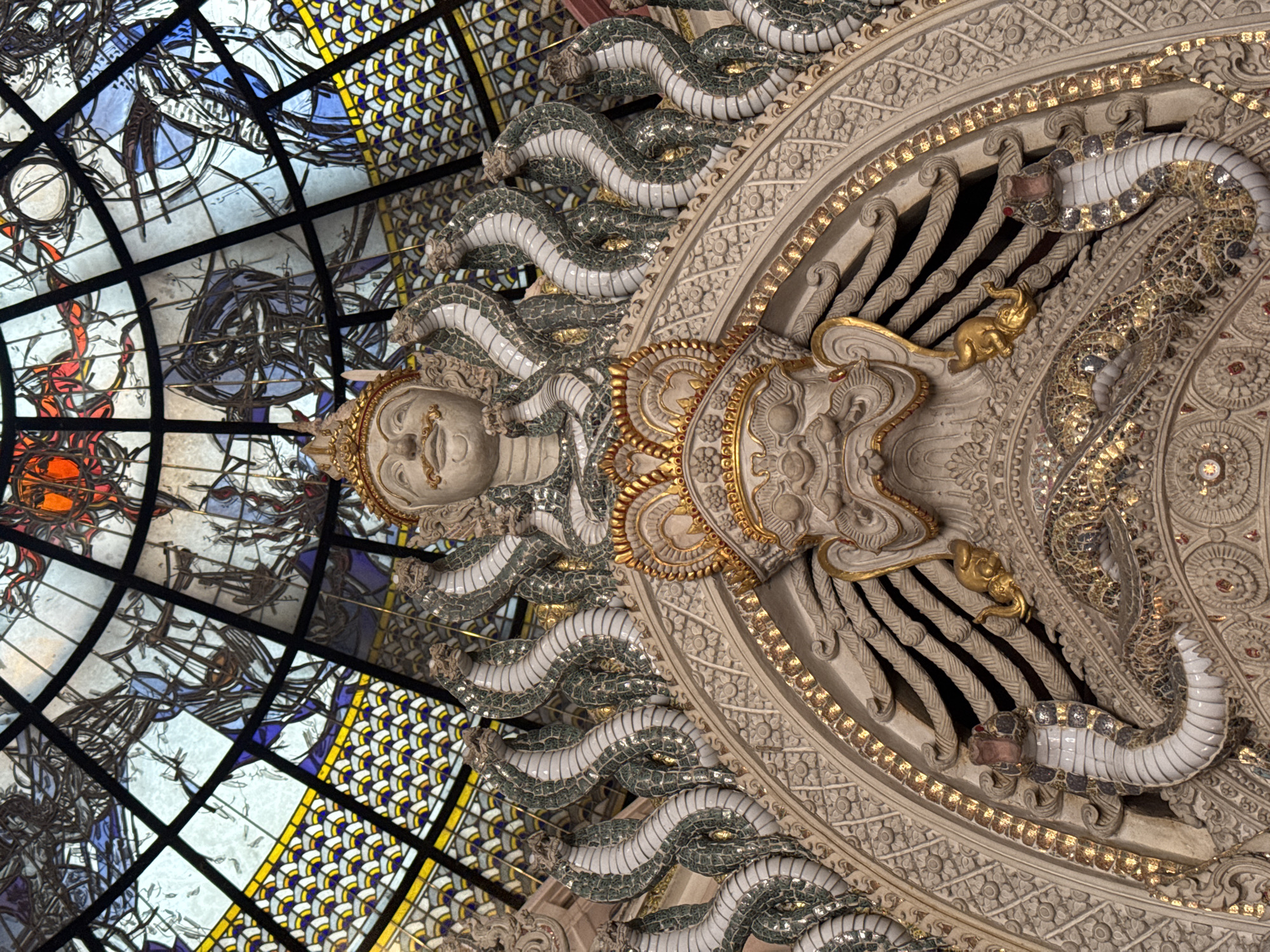 The image shows a close-up view of intricate architectural carvings and a stained-glass ceiling. The carvings depict multiple serpent-like figures with detailed scales and heads, surrounding a central, ornate, crowned face with a mustache. There are also smaller golden elephant figures. The stained-glass ceiling is composed of geometric patterns in yellow, blue, and white, interspersed with darker, abstract designs and splashes of red. The overall impression is of a richly decorated interior space, possibly within a temple or religious building. The provided hints suggest the location is Samrong Nuea Subdistrict Municipality, Thailand, likely within the Erawan Museum, known for its distinctive mythological and artistic elements.