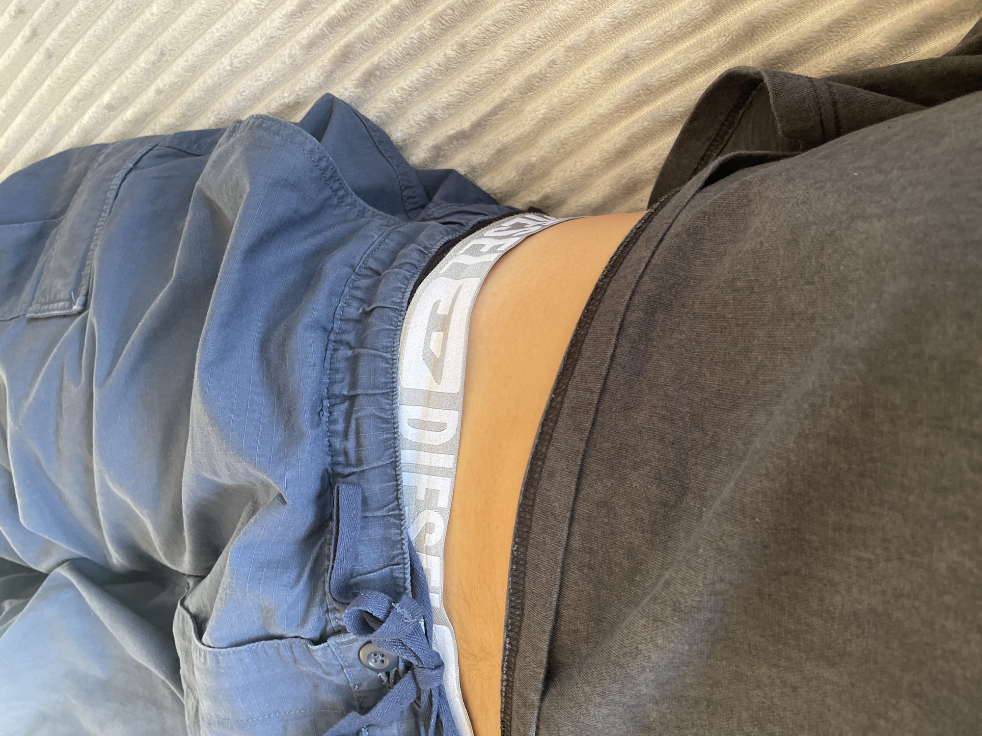 The image shows a person lying on a textured, light-colored surface, likely a bed or sofa. They are wearing blue cargo pants with a buttoned pocket and drawstrings. A grey t-shirt or sweatshirt is partially visible. The waistband of white underwear with the brand name "DIESEL" is showing between the pants and the shirt. There is no indication of the location being in Sestroretsk, Russia.