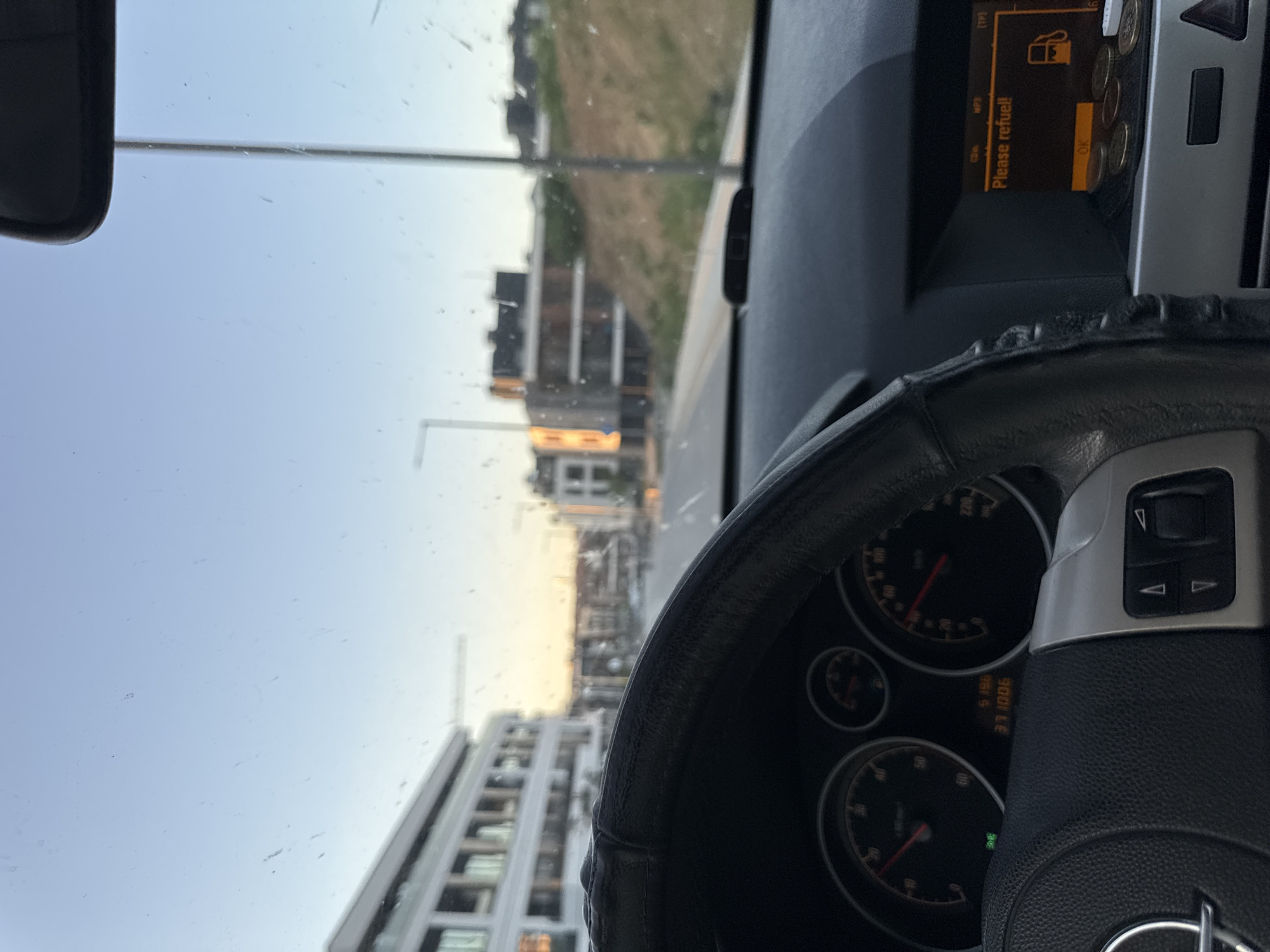 The image shows a view from inside a car, looking out through the windshield. In the foreground, there's a steering wheel with a black cover. The dashboard is visible, displaying a fuel indicator with a "Please refuel!" message. The car is presumably driving through a modern urban area. Outside, buildings are visible, including what appear to be residential or office structures. Based on the style of buildings and overall environment, the location is likely Sofia, Bulgaria. There are no people visible in the image. The focus is on the driving perspective and the car's internal features in relation to the external urban landscape.
