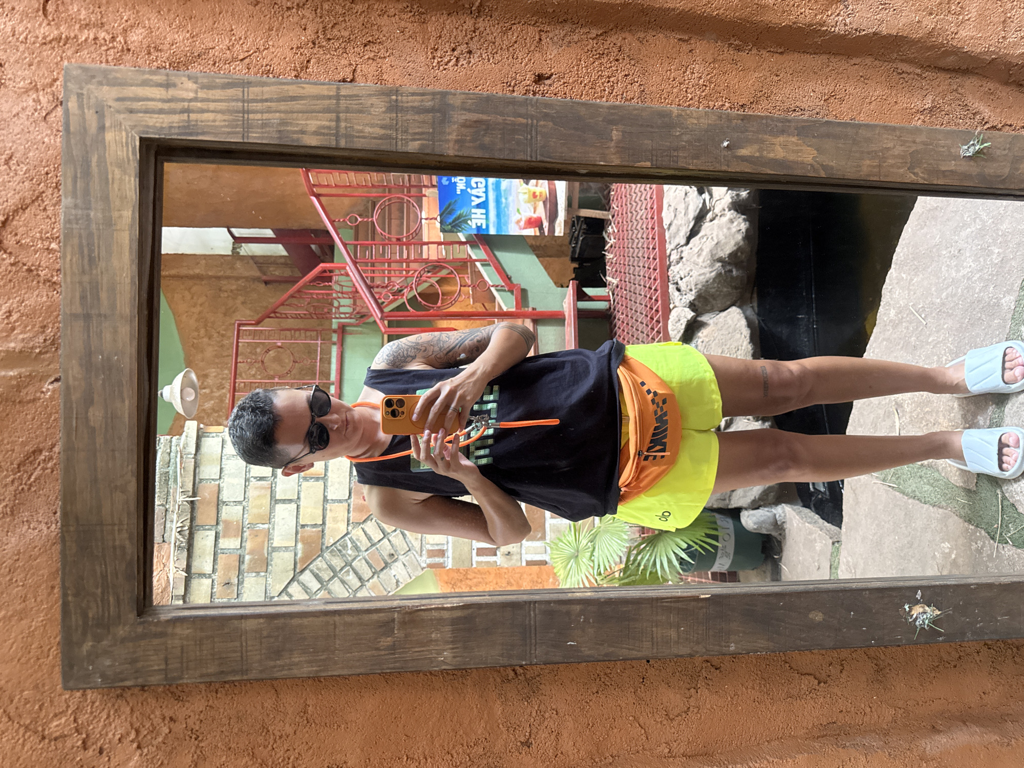 A person stands in front of a mirror, taking a selfie. The person is wearing sunglasses, a black tank top, bright yellow shorts, and white slides. They are holding an orange smartphone. The mirror is framed by a rustic wooden border, and the wall behind it is textured and orange. In the reflection of the mirror, a staircase with red railings is visible, as well as brickwork, plants, and a sign with an image of the beach and drinks. The scene appears to be outdoors, possibly in a courtyard or patio area.