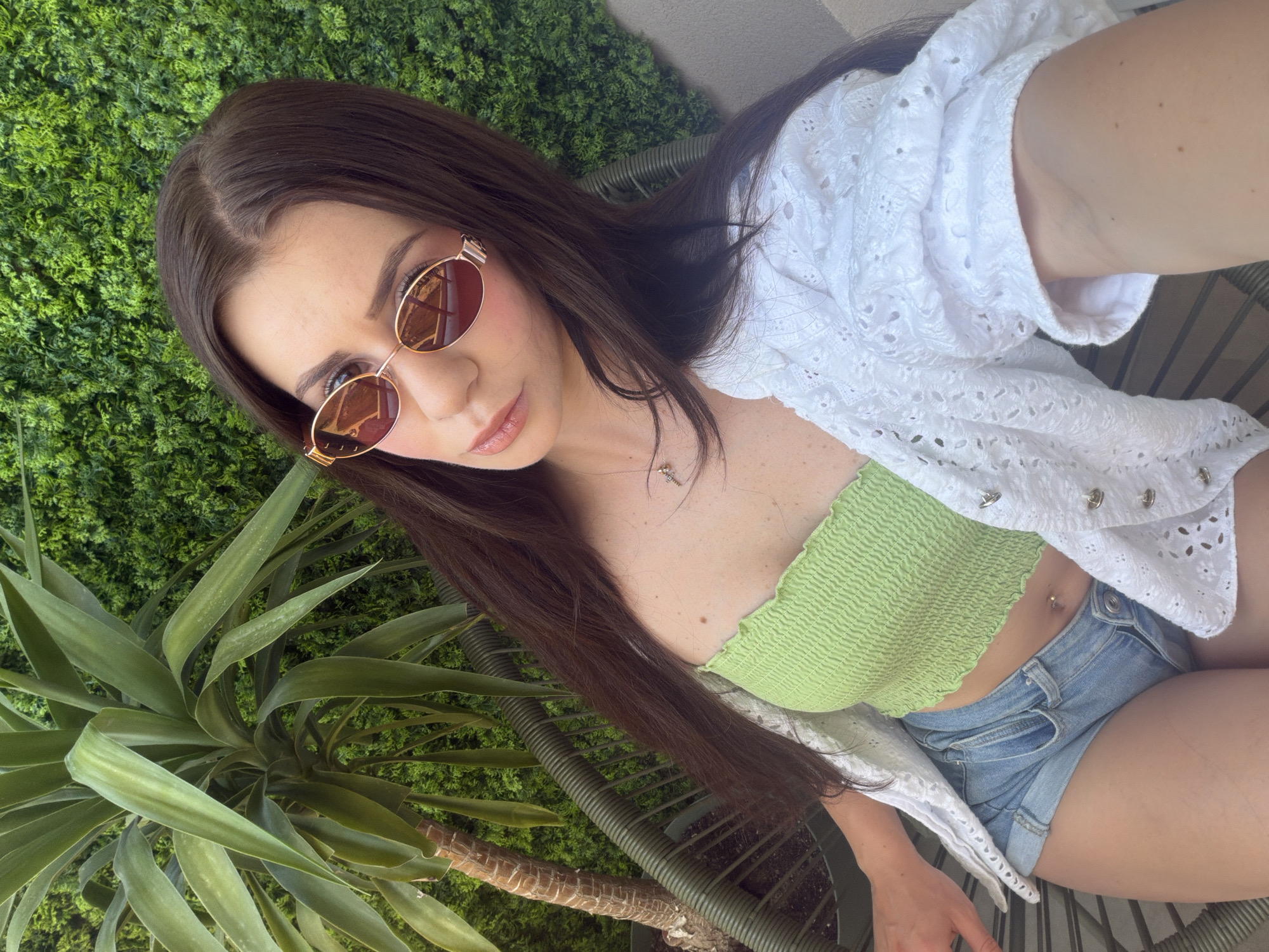A medium, eye-level shot captures a woman sitting in an outdoor setting, possibly in Plovdiv, Bulgaria. The foreground is marked by the presence of a large, green yucca plant. Behind the woman, there is a green wall covered with a dense, foliage-like material.
The woman is dressed in a lime-green tube top, denim shorts, and a white, lacy cover-up. She is wearing rose-tinted sunglasses. The combination of these elements suggests a relaxed, summery atmosphere. The details in the image are clear and distinct, providing a comprehensive view of the scene.
