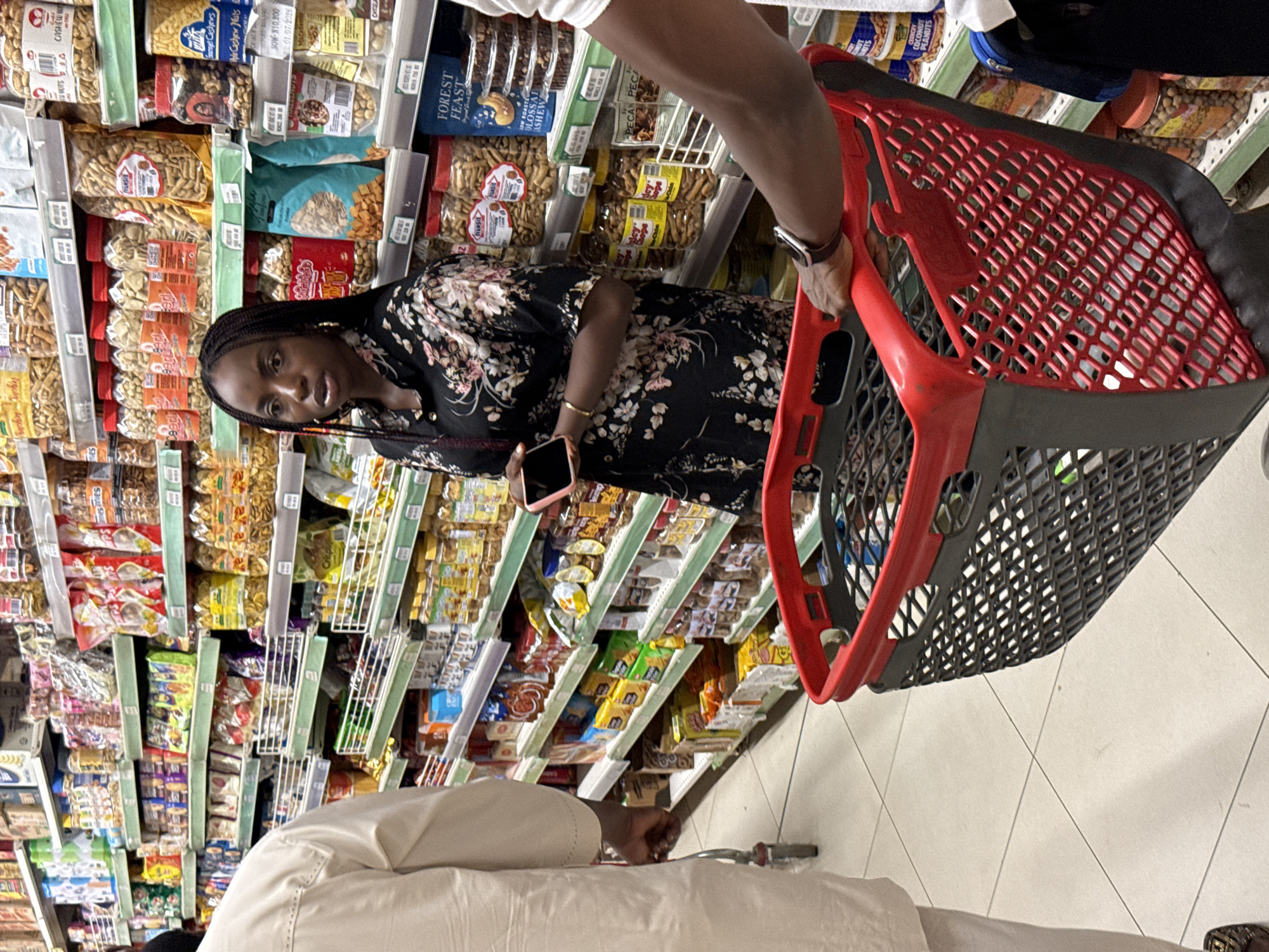 The image depicts a woman in a black floral dress holding a smartphone in a supermarket aisle. She stands near shelves stocked with various nuts and snacks. A red shopping basket is being pushed by someone to her right, and another person in a cream-colored outfit stands to her left. The location is implied to be a supermarket in Abuja, Nigeria, based on the text and the setting.