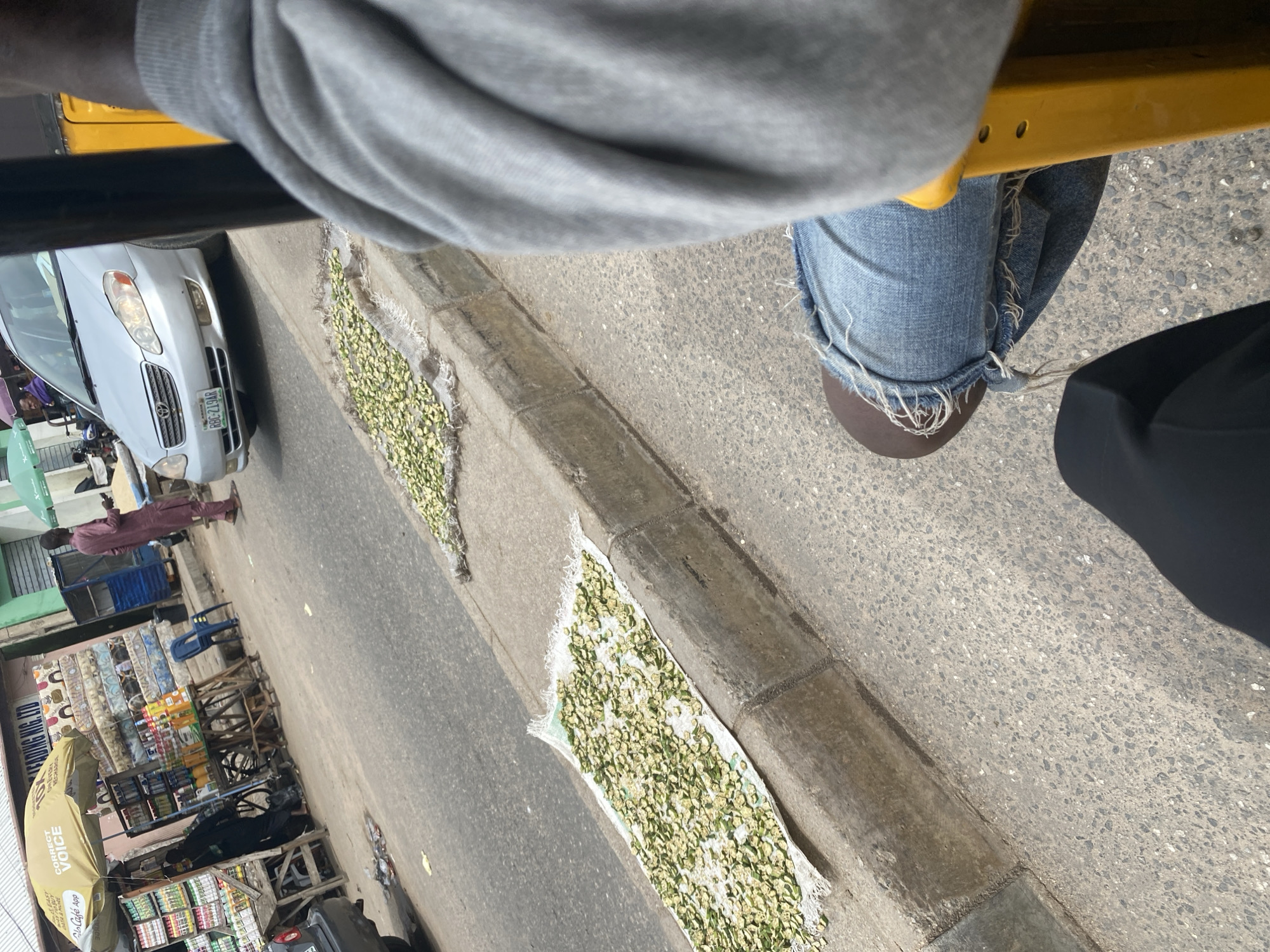 The image, taken in Jalingo, Nigeria, depicts an outdoor scene, likely a street or roadside market. The perspective is from inside a vehicle, looking outwards and slightly downwards. In the foreground, we see the legs of a person wearing blue jeans with frayed hems and brown shoes, and a grey hooded garment. The person appears to be a passenger.
Outside the vehicle, the immediate focus is on a row of what appear to be food items, possibly okra or a similar vegetable, laid out to dry on white cloths along the edge of the road, next to a concrete curb. A silver Toyota sedan is parked to the left, with its headlights visible. Further down the street, a small market stall is set up with various goods displayed on shelves and tables, including drinks and other items. A person in a maroon outfit is standing near the market stall, looking towards the left. A blue plastic chair is also visible near the stall. An yellow umbrella with text is partially visible. The overall impression is of a busy street scene in a sunny climate. The time of day appears to be daytime, with bright light casting shadows.