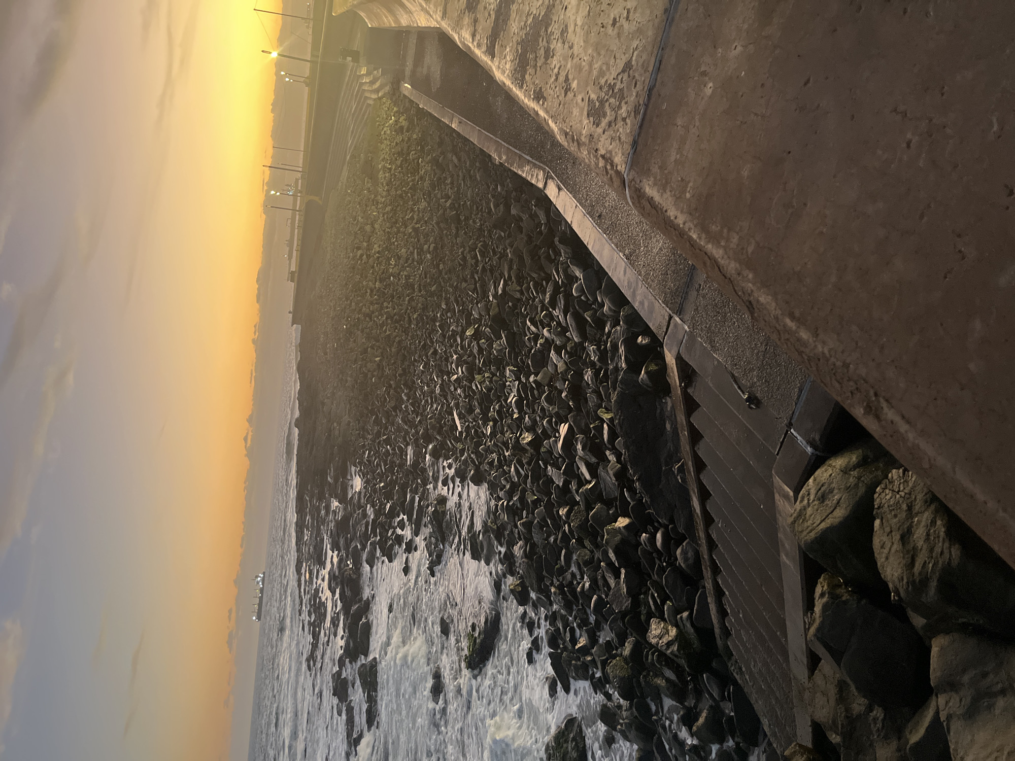 This image, taken during sunset in Arica, Chile, shows a rocky shoreline adjacent to a concrete seawall. The water is calm and reflects the warm colors of the setting sun. A long ship is visible in the distance. The foreground consists of large, dark rocks interspersed with crashing waves. The seawall has built-in steps. There are lampposts on the horizon.