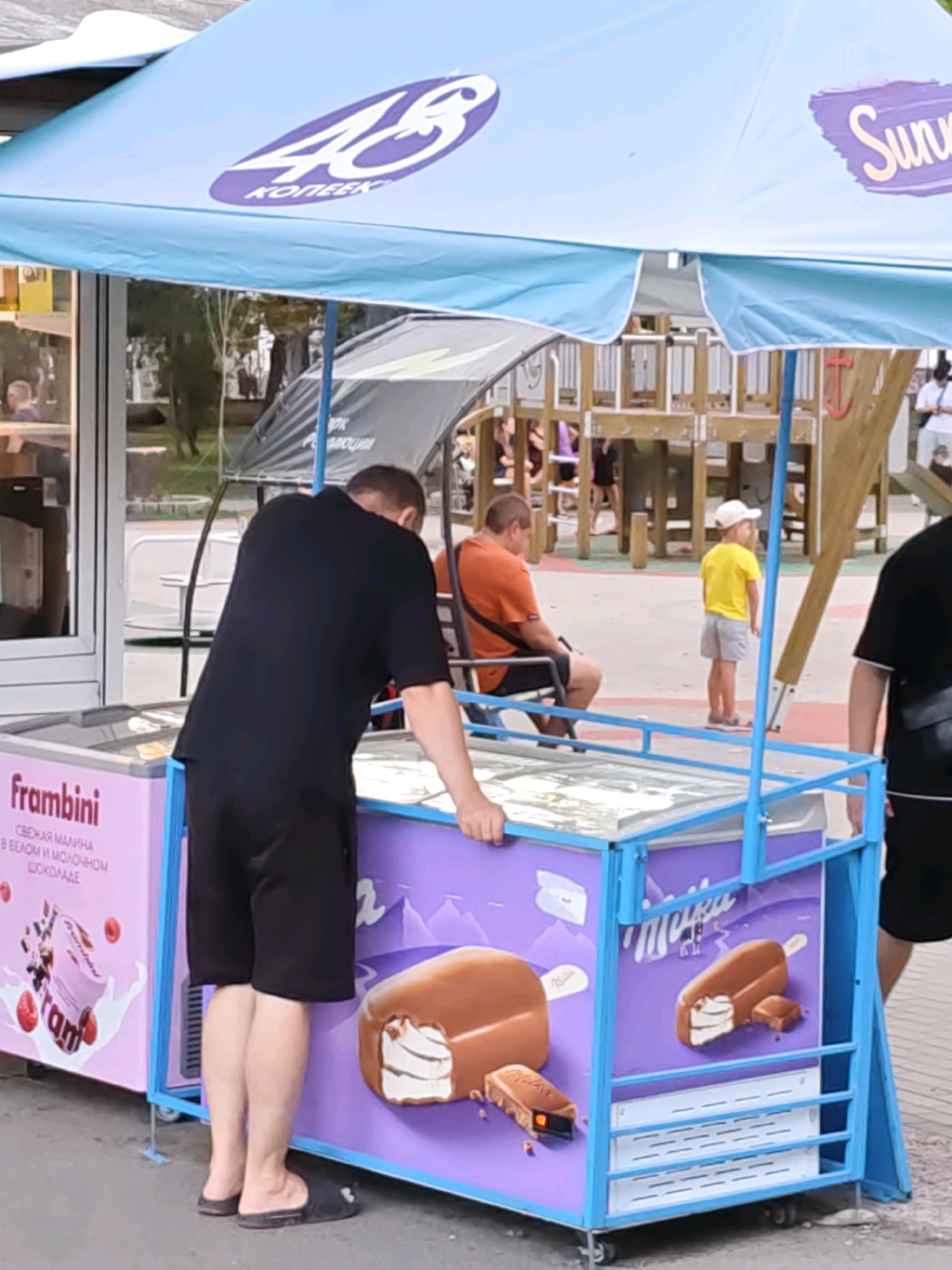 An outdoor scene captures an ice cream stand with a light blue awning. A man in a black t-shirt and black shorts stands in front of an ice cream freezer, bending over and reaching into it. Another person is partially visible on the far right, wearing a black t-shirt. In the background, a person in an orange t-shirt sits on a bench, and a child in a yellow t-shirt and grey shorts stands near a wooden playground structure. The ice cream freezer displays images of ice cream bars and has text on its side. The ground appears to be pavement. The scene suggests a public area, possibly a park or recreational space in the city of Rostov-on-Don, Russia.