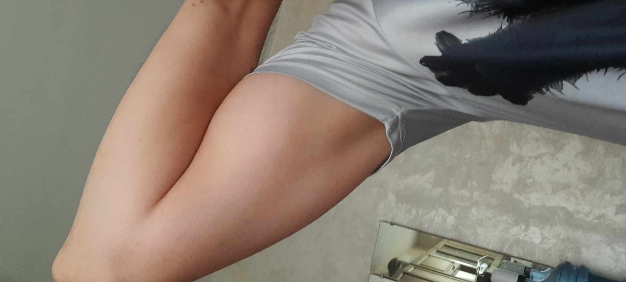 A close-up, angled shot focuses on a person's upper thighs and torso, wearing a light gray t-shirt with a black graphic design of an animal's head and mane. The background features a textured wall with a light, marbled pattern. To the left, a partial reflection in a mirror shows a cabinet with glass doors and a person's arm holding a blue object. The lighting suggests an indoor environment.