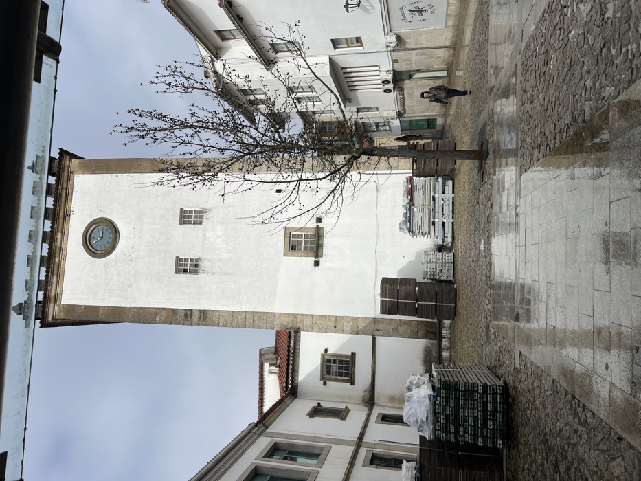 The scene depicts an urban courtyard in Bragança, Portugal, under an overcast sky, with the ground visibly wet and reflective. A tall, rectangular clock tower, primarily white with exposed stone quoins at its corners, dominates the central background. Near its apex, a circular clock face displays approximately 11:10. Below the clock, two small, square, gridded windows are visible, with a larger rectangular gridded window positioned further down. The tower is crowned with crenellated battlements.
To the immediate left of the tower, a white building with a reddish-brown tiled roof extends, showing multiple rectangular windows with stone sills. On the right, another multi-story white building with several windows and visible exterior air conditioning units lines the paved area.
The ground is composed of a mix of cobblestones and larger grey flagstones, uniformly wet from recent precipitation, reflecting ambient light. In the mid-ground, at the base of the tower and extending to the left, are stacks of materials. These include pallets of green-banded items, some covered in white plastic sheeting, and stacks of dark wooden crates. To the right of these, lighter-colored, possibly metal, frames or panels are stacked.
A single leafless deciduous tree with visible dark buds or berries stands in the mid-ground, positioned between the tower and the building on the right.
One individual is present, walking from the mid-ground towards the background on the right side of the paved area. This person is dressed in dark clothing, including a hooded jacket or coat, and dark trousers. Their posture indicates forward motion. No other individuals or direct interactions are visible.