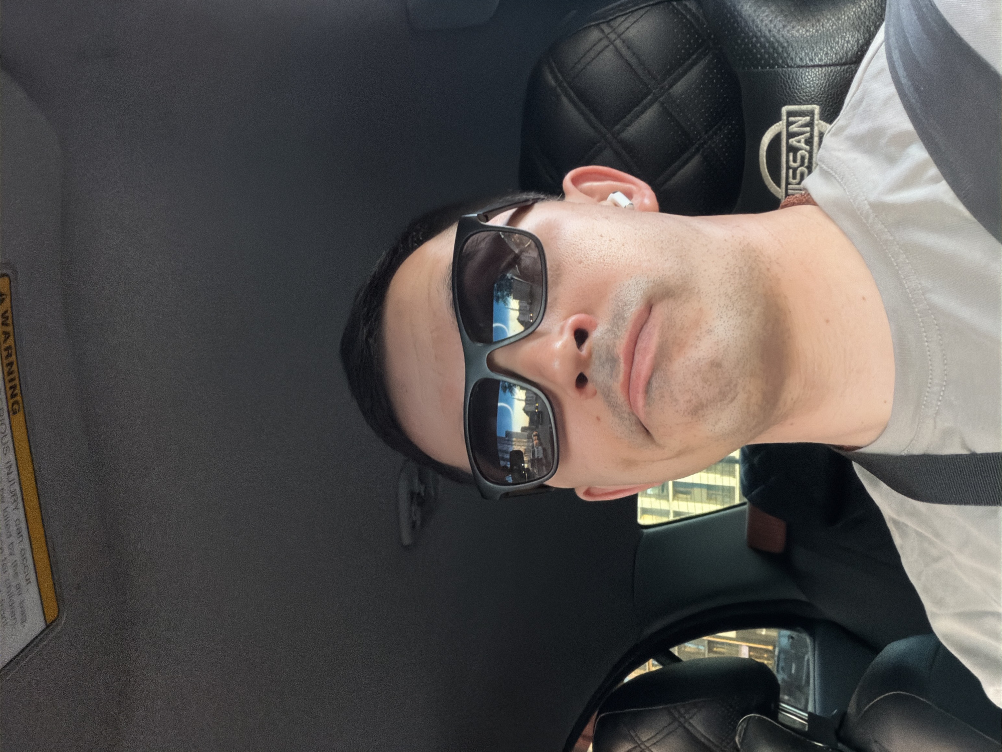 A man wearing sunglasses sits in the driver's seat of a Nissan car. The car interior is mostly black with a quilted pattern on the headrest. A Nissan logo is visible on the headrest. The man has an earbud in his left ear and is wearing a light gray shirt with a gray strap or seatbelt across his shoulder. Reflected in the sunglasses are buildings that appear to be in a city environment. The image suggests the man is either driving or stopped in traffic. A warning label is visible on the car's sun visor. No specific features in the reflection of the glasses or the surroundings indicate Izhevsk, Russia.
