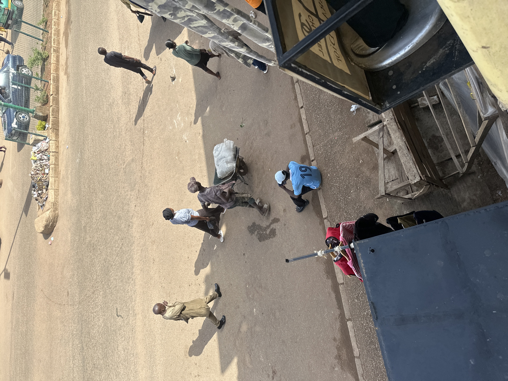 An overhead, medium shot captures a street scene in Bukuru, Nigeria. Several individuals are present on a paved road. A man in a tan robe walks from left to right, looking down. To his right, a woman in a light blue top and dark skirt walks away from the viewer. Further right, a man in a grey shirt and pants pushes a wheelbarrow loaded with a large white sack. To the right of him, a man in a green top and dark shorts walks away from the viewer. In the lower right of the image, a young person wearing a light blue shirt with the number "19" on the back sits on a curb, looking down. A wheeled cart with various items of clothing, including a red garment, is parked on the curb to the left of the seated person. The environment consists of a paved road and a curb, with shadows cast by the sun. In the background, a dark blue car and a green vehicle are visible, along with a pile of debris. The scene appears to be taking place in daylight.