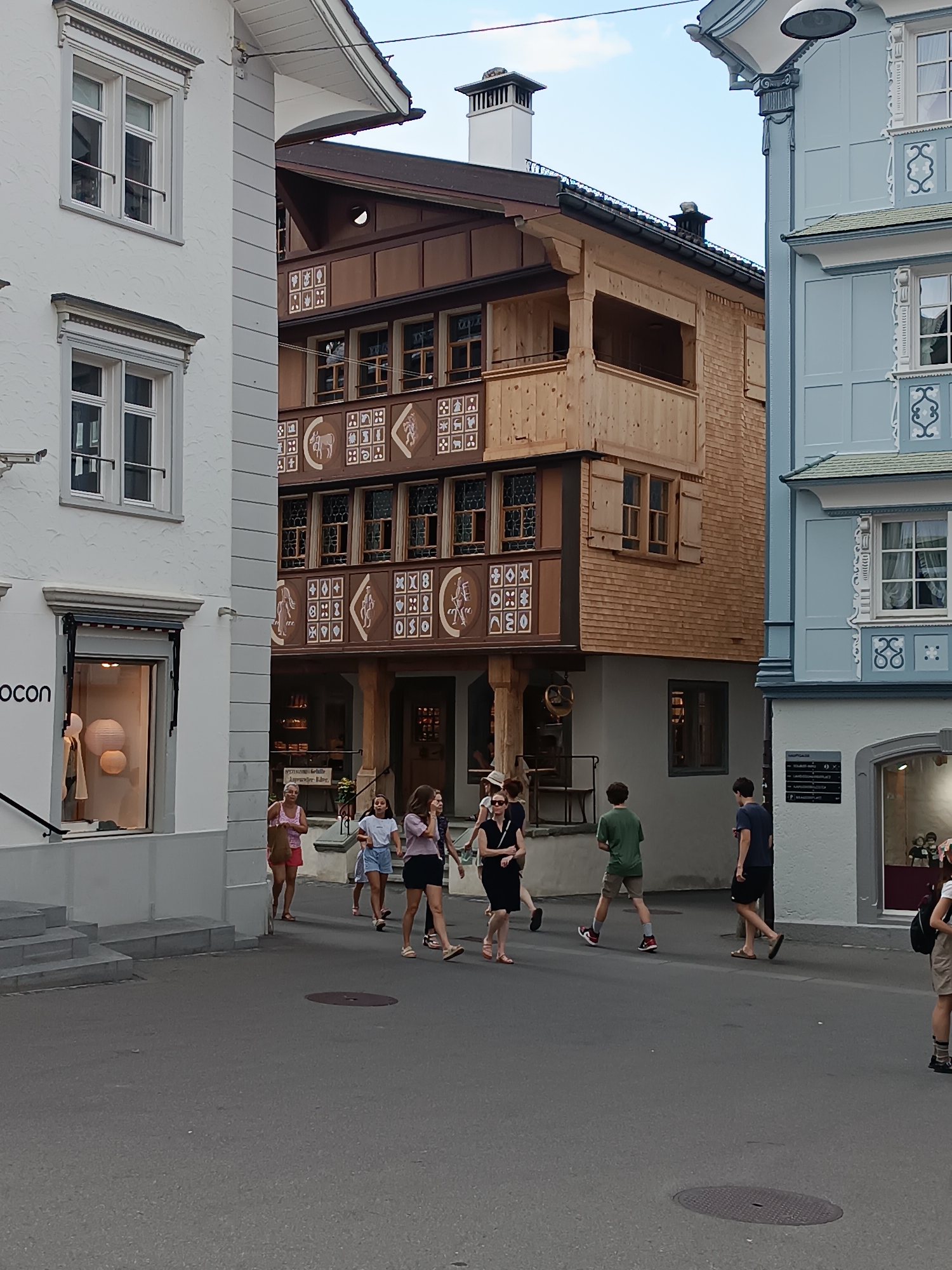 In Appenzell, Switzerland, a street scene unfolds with architectural diversity. On the left, a white building features two rows of windows, each with decorative trim. Adjacent to it, a traditional wooden building with dark brown trim showcases intricate decorative panels depicting geometric patterns and animal figures, topped by a gabled roof. To the right, a pale blue building with white accents and shuttered windows adds another layer of architectural style. The street is paved, and several people are walking. A group of women and girls are moving from left to right, some carrying bags. Two young men are walking in the same direction on the right side of the street. The lighting suggests it is daytime.