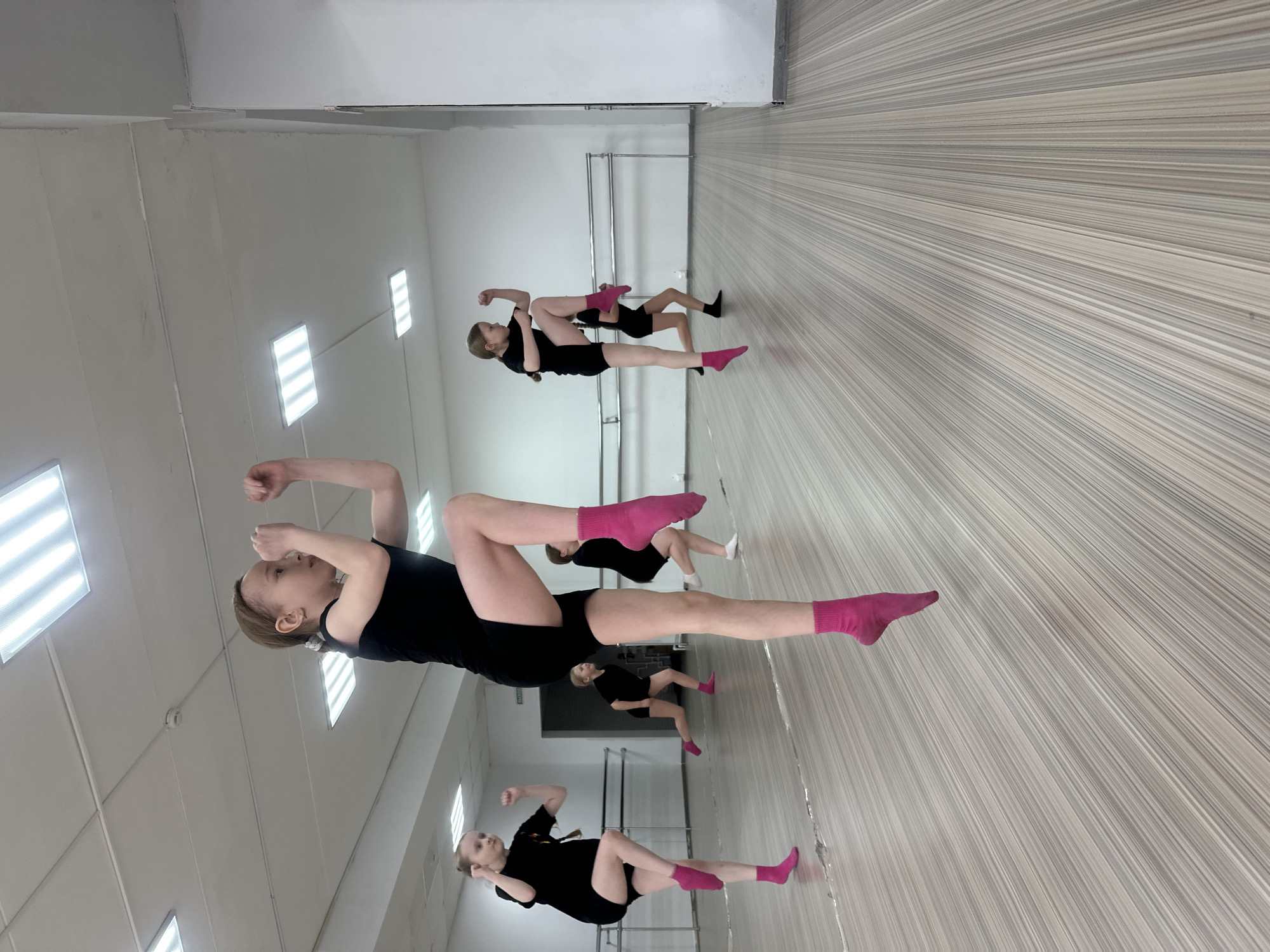 An image captures a dance or gymnastics class taking place indoors, likely in a studio or gymnasium in Votkinsk, Russia. The scene features multiple young individuals, primarily girls, dressed in black athletic wear. The most prominent figure, a girl with light brown hair, is in the foreground, striking a pose with her arms bent and raised, and her knees slightly bent. She wears black shorts and bright pink socks.
The studio has a large mirrored wall on the right, reflecting other participants and elements of the room. In the reflections, several other girls are visible, some in similar poses and others appearing to be stretching or standing. Most of them also wear black attire and brightly colored socks, with pink being common, though one reflection shows a person in white socks and another in black socks. A metal barre is visible along the mirrored wall.
The studio features a light-colored, striped wooden floor and white walls. The ceiling is also white, equipped with multiple rectangular fluorescent light fixtures, providing bright, artificial illumination. Above one of the reflected figures, the Russian word "ВЫХОД" (EXIT) is visible on the wall, indicating an exit sign. The atmosphere appears focused, with the participants engaged in their physical activity.
