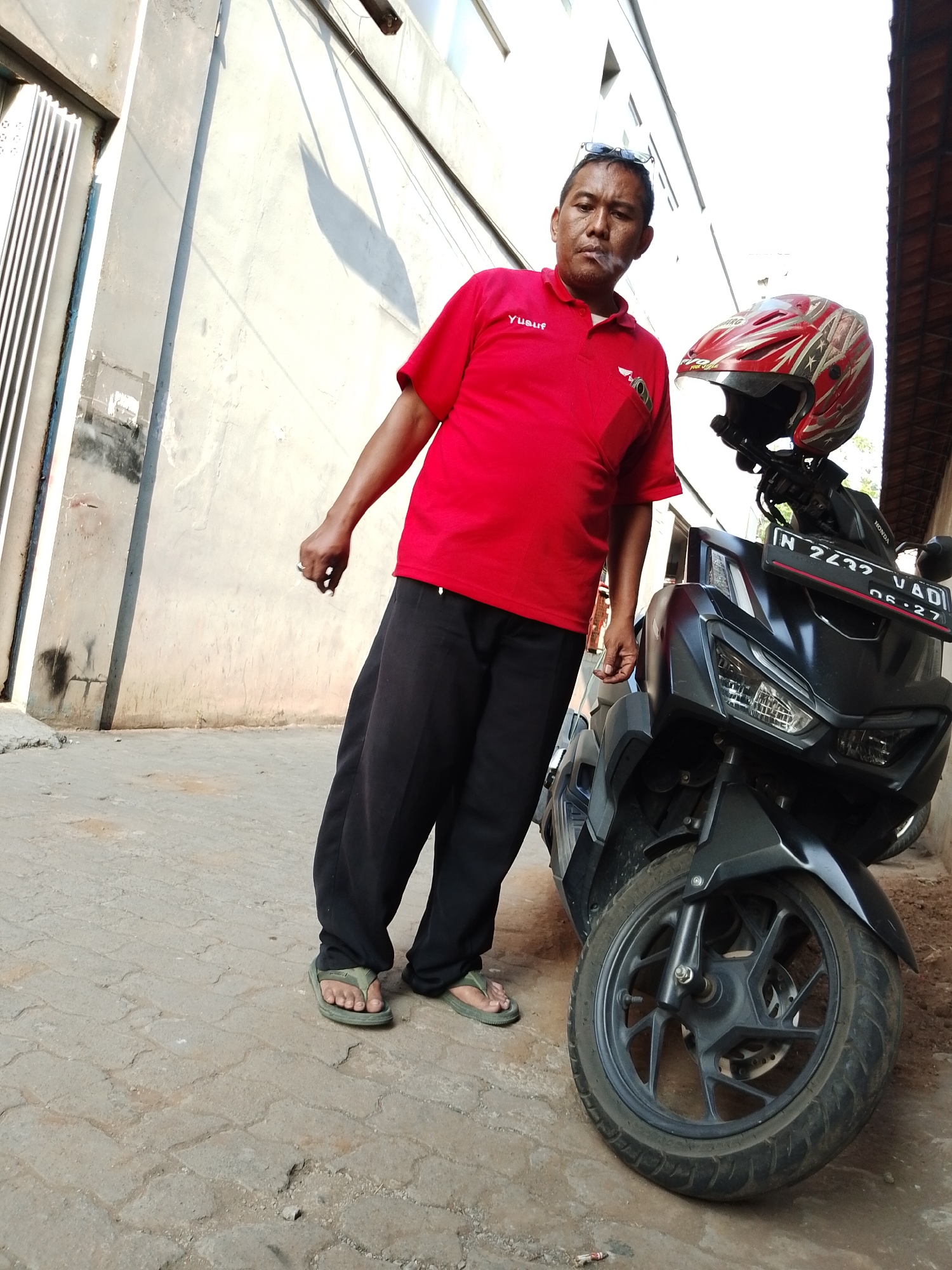 A man of Asian descent, appearing to be in his late 40s to early 50s, stands next to a black motorcycle. He is wearing a red polo shirt with the name "Yusuf" embroidered on the left chest, black trousers, and green flip-flops. He is smoking a cigarette. The motorcycle has a red and white helmet with a star design resting on its handlebars. The license plate reads "N 2422 VAD" and "06-27". The background consists of a light-colored concrete wall, a metal gate, and a tiled roof. The scene is outdoors on a paved surface, suggesting a location such as a street or alleyway. The lighting indicates it is daytime, with shadows cast by the building and the man. The overall impression is a candid, everyday moment. The location is identified as Pasuruan, Indonesia.