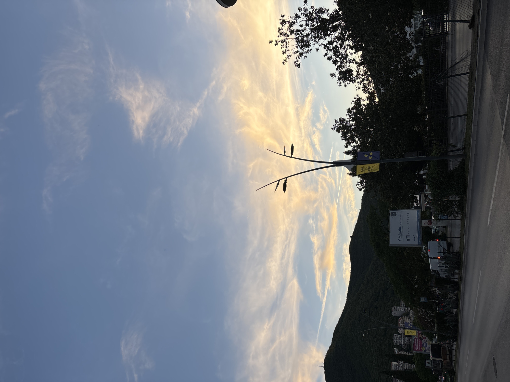 The image depicts a street scene in Budva, Montenegro, during what appears to be dusk or early evening. The sky is a mix of blue and pale yellow, with wispy clouds that catch the last rays of sunlight. In the foreground, a paved road runs horizontally across the frame. A white van is parked on the right side of the road, and further down the road, there are indications of traffic.
To the left of the road, a dark green, tree-covered hill dominates the background, with buildings built into its slope. A large billboard advertising "CMM CHESTERTONS N°1 REAL ESTATE" is prominently displayed on the right side of the road, behind a dark metal fence. A modern street lamp with multiple arms and light fixtures stands near the billboard. Two flags, one yellow and one blue, are attached to the street lamp pole. Silhouettes of trees are visible against the sky and the hill. The overall impression is of a city street transitioning from day to night, with natural elements and urban infrastructure coexisting.