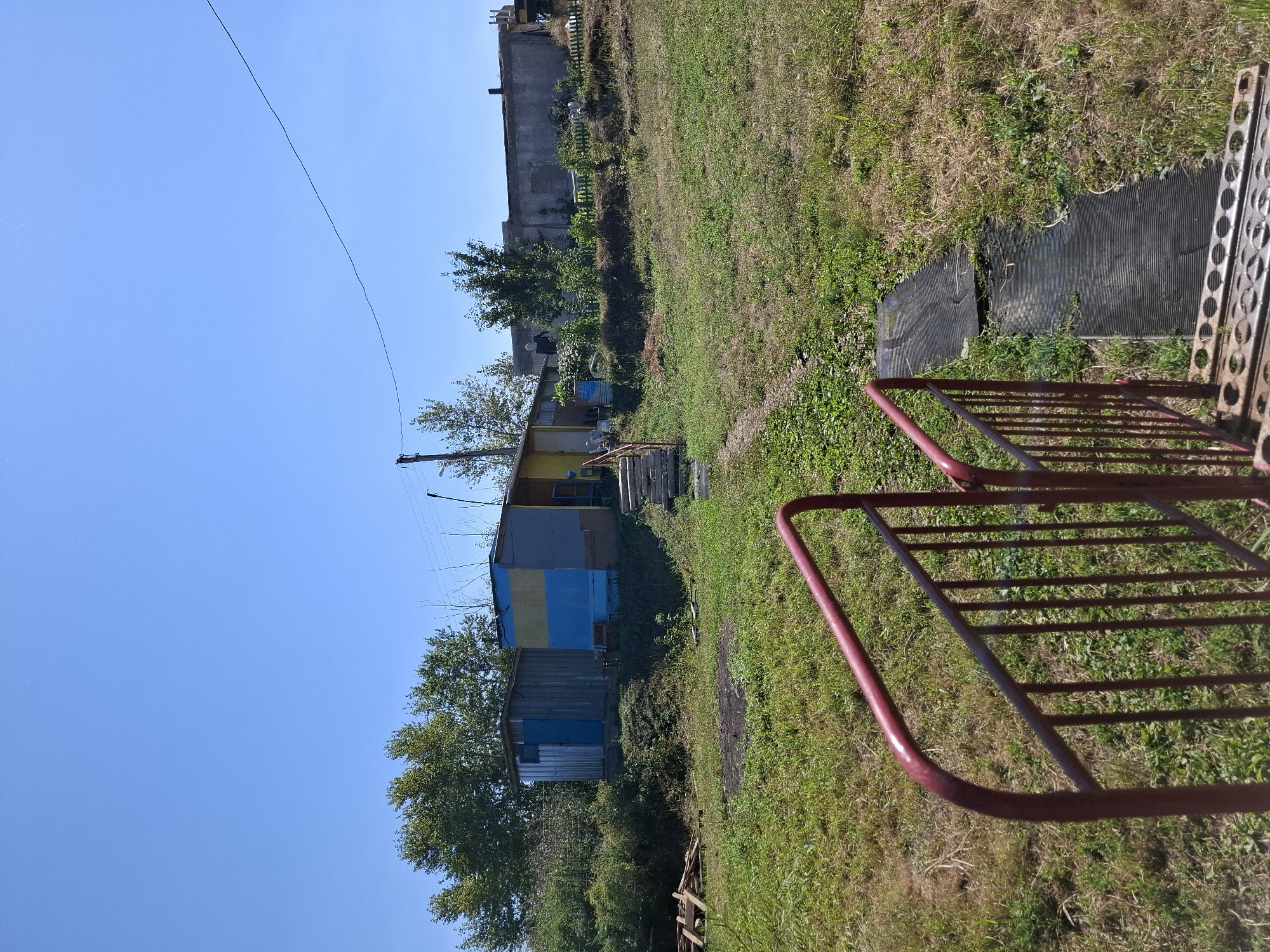 An exterior, low-angle, wide shot captures a bright, sunny day with a clear blue sky above. In the foreground, a section of a red metal railing is visible, leading the eye towards a weathered, dark-colored ramp or walkway on the ground. Beyond the ramp, a path winds uphill towards a cluster of small, brightly colored structures. One structure features blue and yellow siding, while another adjacent one has a silver, corrugated metal exterior with a blue door. Trees with green foliage line the background, and a long, grey, industrial-looking building is situated further back. The ground is covered in dry, green grass and patches of dark earth.