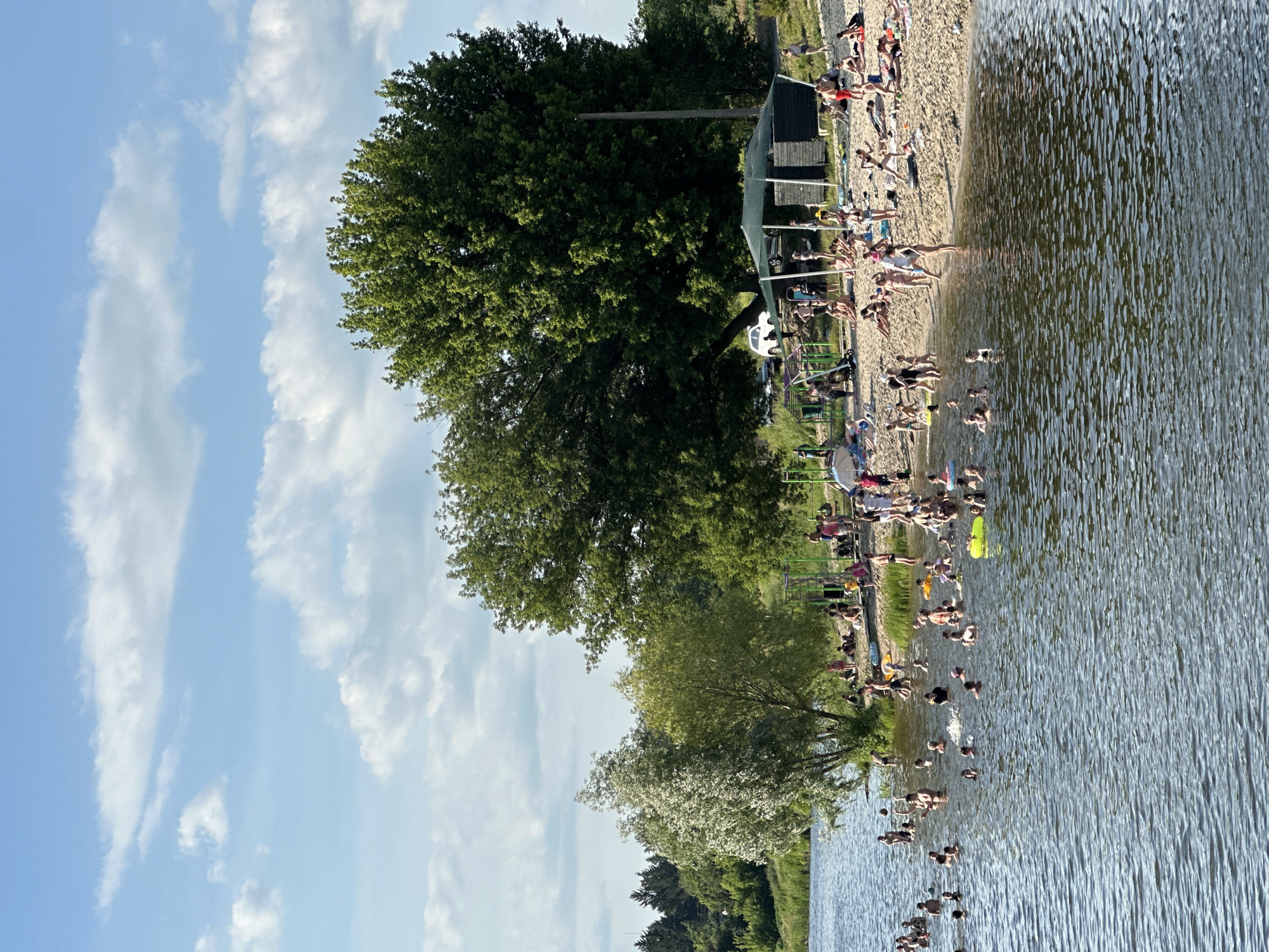 The image shows a crowded beach scene with many people enjoying a sunny day at the water. The perspective is from slightly above and to the side, looking down towards the beach and the water. The sky is blue with scattered white clouds. There are large green trees along the edge of the beach and in the background.
On the sandy beach, people are sunbathing, sitting, and interacting. Some are wearing swimwear. There are some beach chairs and umbrellas. Further back, there's a structure that appears to be a changing room or small building, and some playground equipment.
In the water, many people are swimming, wading, and playing. The water is a calm blue with ripples from the activity. A bright yellow inflatable float is visible near the shore.
The scene appears to be a popular recreational area, likely a lake or a calm bay, given the environment. The bright sunlight suggests it is daytime, likely midday or afternoon, and the weather is warm and pleasant, encouraging outdoor activities. The overall mood conveyed is one of relaxation and enjoyment on a summer day. There is no visible text in the image.
