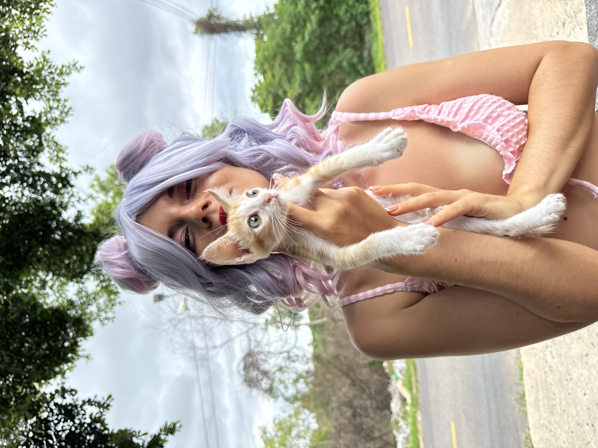 A person with light purple and pink hair, styled with two buns on top, holds a small, light orange and white kitten in front of their face. The person is wearing a pink gingham bikini top. They are outdoors, with green trees and a cloudy sky in the background. A road is visible to the right of the person. The scene appears to be in Mũi Né, Vietnam, suggesting a relaxed, outdoor setting.