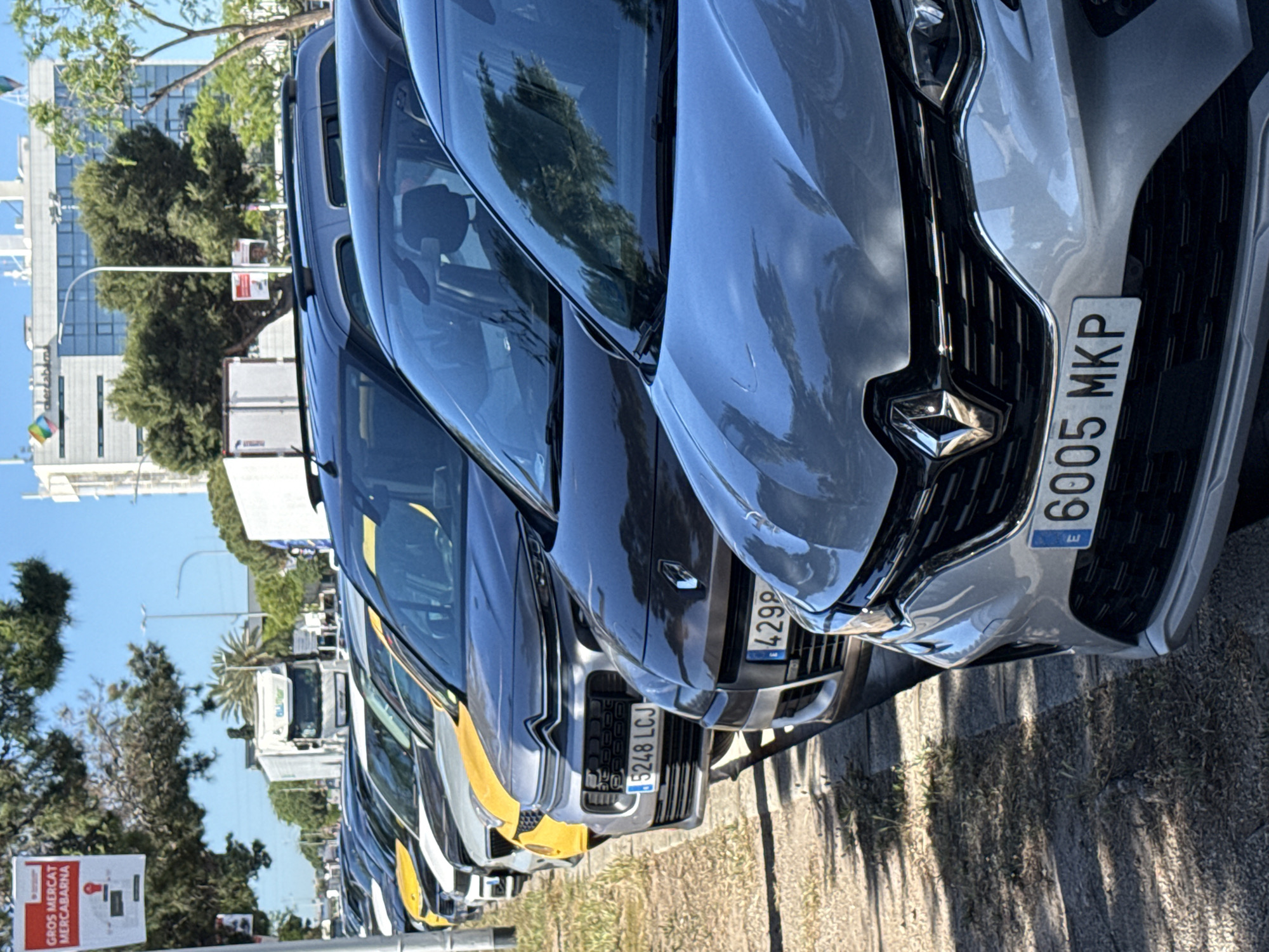 A line of parked Renault cars, partially obscured from one another, is arranged along a roadside in Barcelona, Spain. The cars vary in color, with one car exhibiting a yellow and white scheme. Each car displays a visible license plate, including “6005 MKP,” “4299,” and “5248 LCJ.” The environment consists of trees, a light-colored building, and street signs. The light suggests it is daytime.