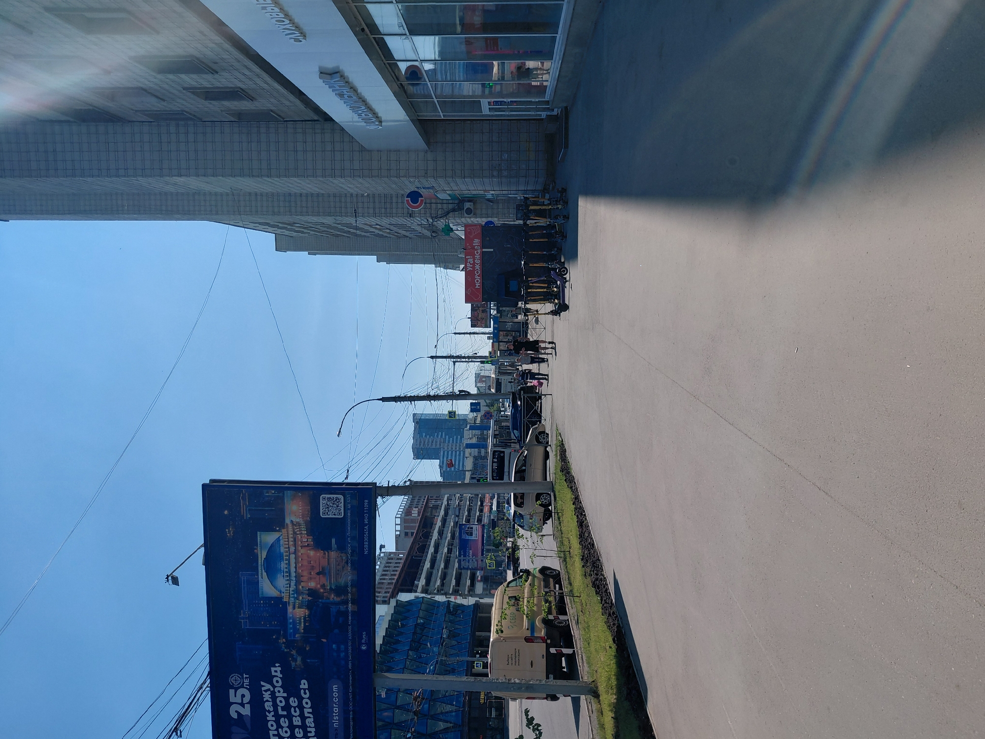 The image depicts a street scene in Novosibirsk, Russia, on a clear day. In the foreground, a gray asphalt sidewalk runs alongside a road. To the left, a grassy area is visible, bordered by a curb. Across the road, a billboard with the text "25 лет" and an image of a building with a dome stands prominently. Further down the street, several buildings line the right side of the road, one of which features large glass windows and signage that appears to be for a bank. A row of electric scooters is parked on the sidewalk in front of a structure with red signage that reads "УРА! МОРОЖЕНОЕ!!!". Several individuals are walking on the sidewalk and crossing the street, and vehicles, including cars, a van, and what appears to be a bus, are present on the road. Power lines crisscross the sky, and a street lamp is visible in the upper right quadrant of the image. The architecture suggests an urban environment.