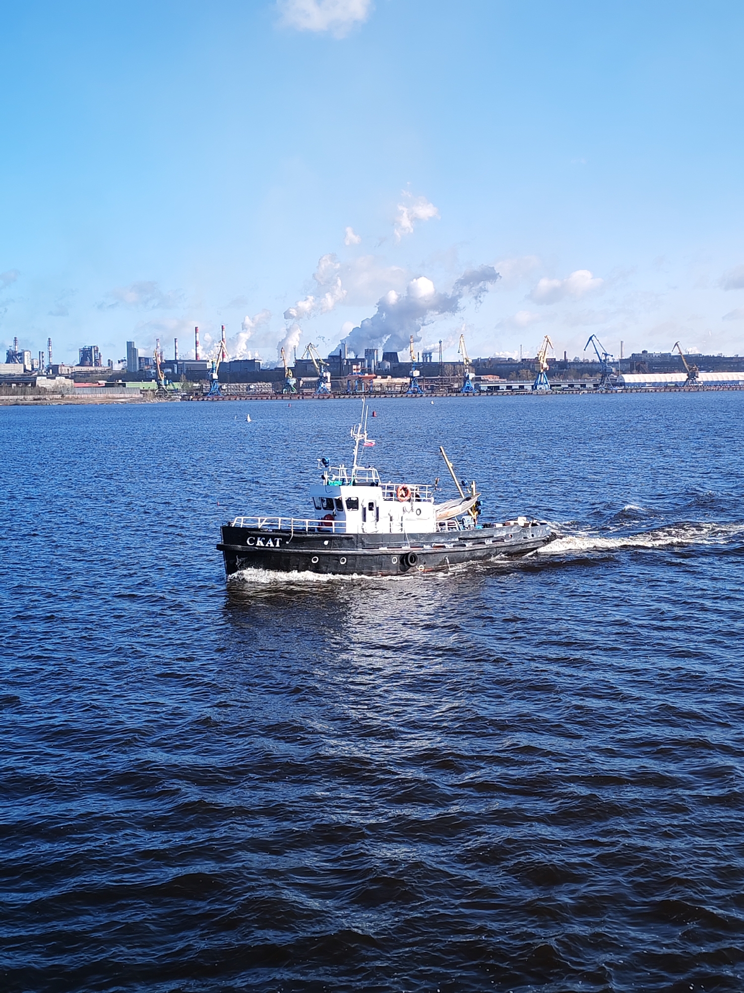 The image shows a black and white tugboat named "CKAT" (SKAT) moving across a body of water, likely the Sheksna River or a similar waterway near Cherepovets, Russia. The water has a choppy surface, indicating some wind. In the background, a cityscape is visible featuring industrial structures, including factories with tall smokestacks and gantry cranes. The smokestacks are emitting plumes of smoke, suggesting active industrial operations. The sky is blue with some scattered clouds.
