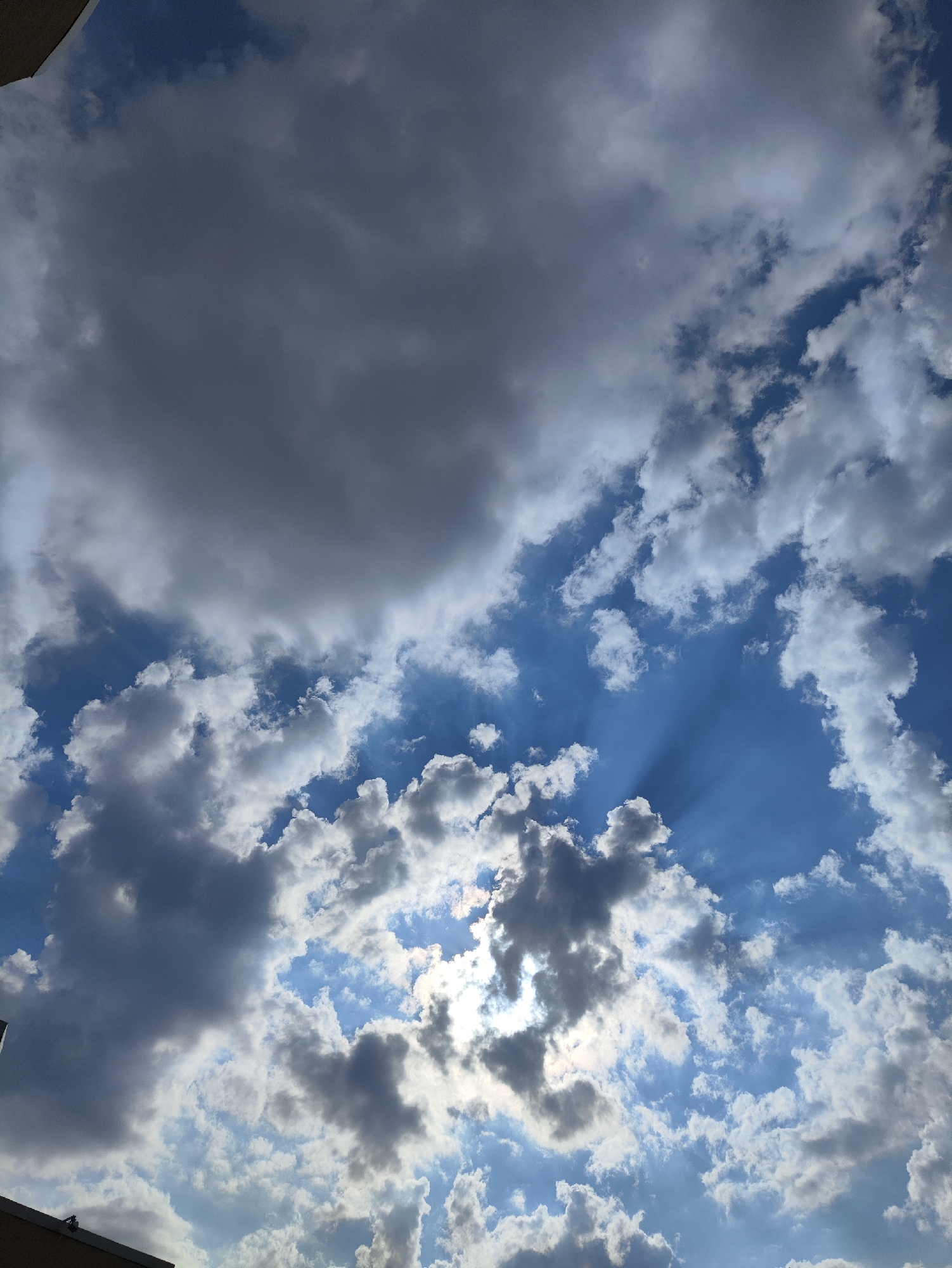 The image captures a dynamic sky filled with a mix of dark, heavy clouds and brighter, scattered cumulus clouds. Sunlight streams through breaks in the clouds, creating visible sunbeams that radiate outwards. The sky is a vibrant blue in the clearer sections. A sliver of a building's edge, possibly a rooftop, is visible in the bottom left corner. This scene is a view of the sky over the city of Kharkiv, Ukraine.
