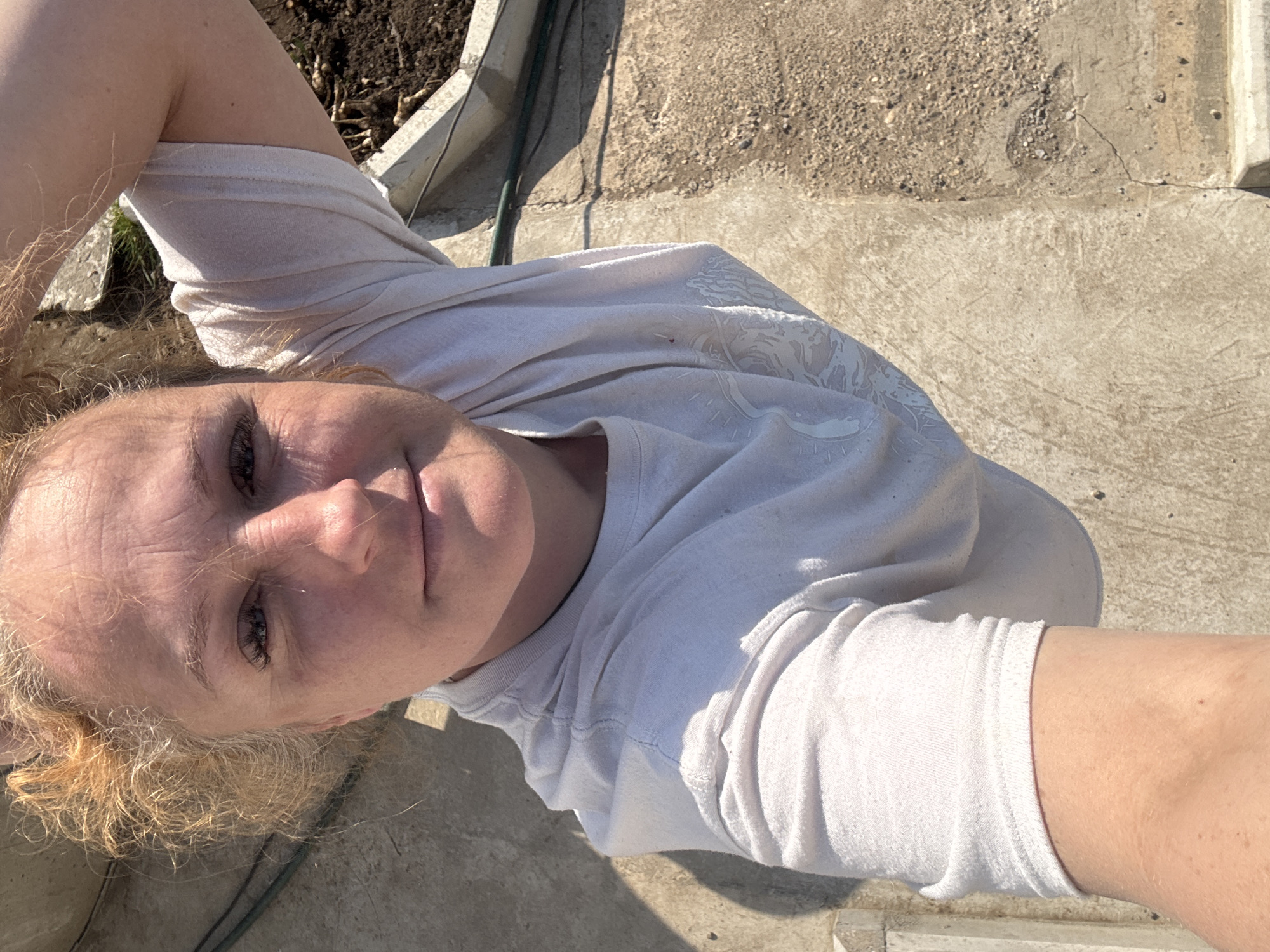 A woman with blond hair and a white t-shirt is lying down on a concrete surface. In the background, there is a planter with soil and some plant matter. The location is likely an outdoor area with concrete paving. There is no conclusive evidence to suggest the location is Chaikovsky, Russia.