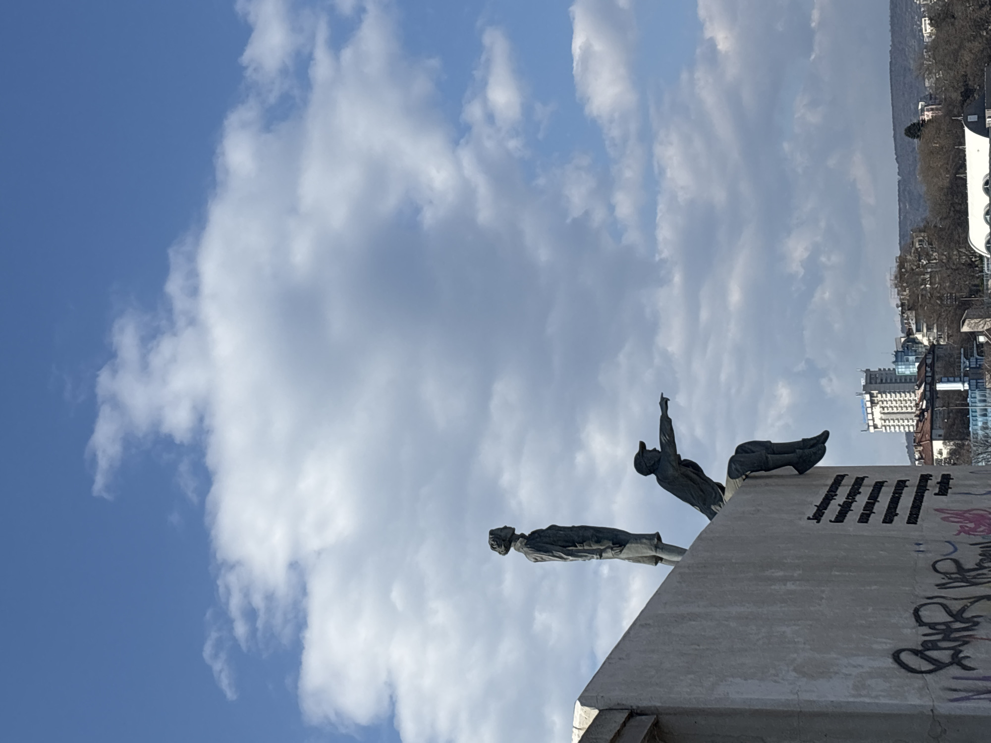 The image depicts the Soviet Army Monument in Varna, Bulgaria. Two bronze statues, one lying down and pointing upwards, the other standing horizontally on a concrete plinth, are prominently featured against a sky with blue patches and cumulus clouds. Graffiti is visible on the concrete plinth. In the background, buildings and trees suggest an urban landscape.